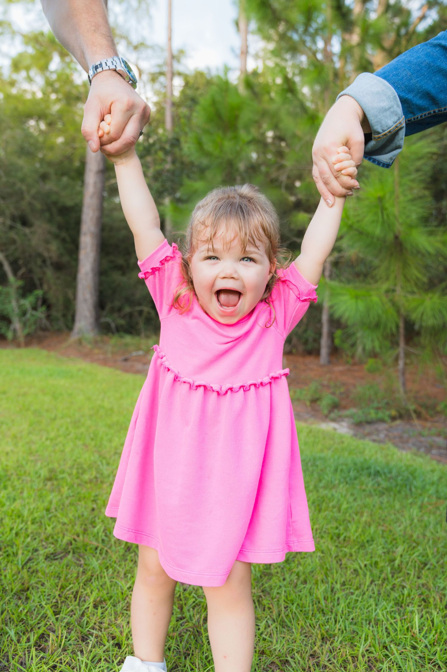 Joyful toddler girl in pink dress laughing while being lifted by both hands outdoors.