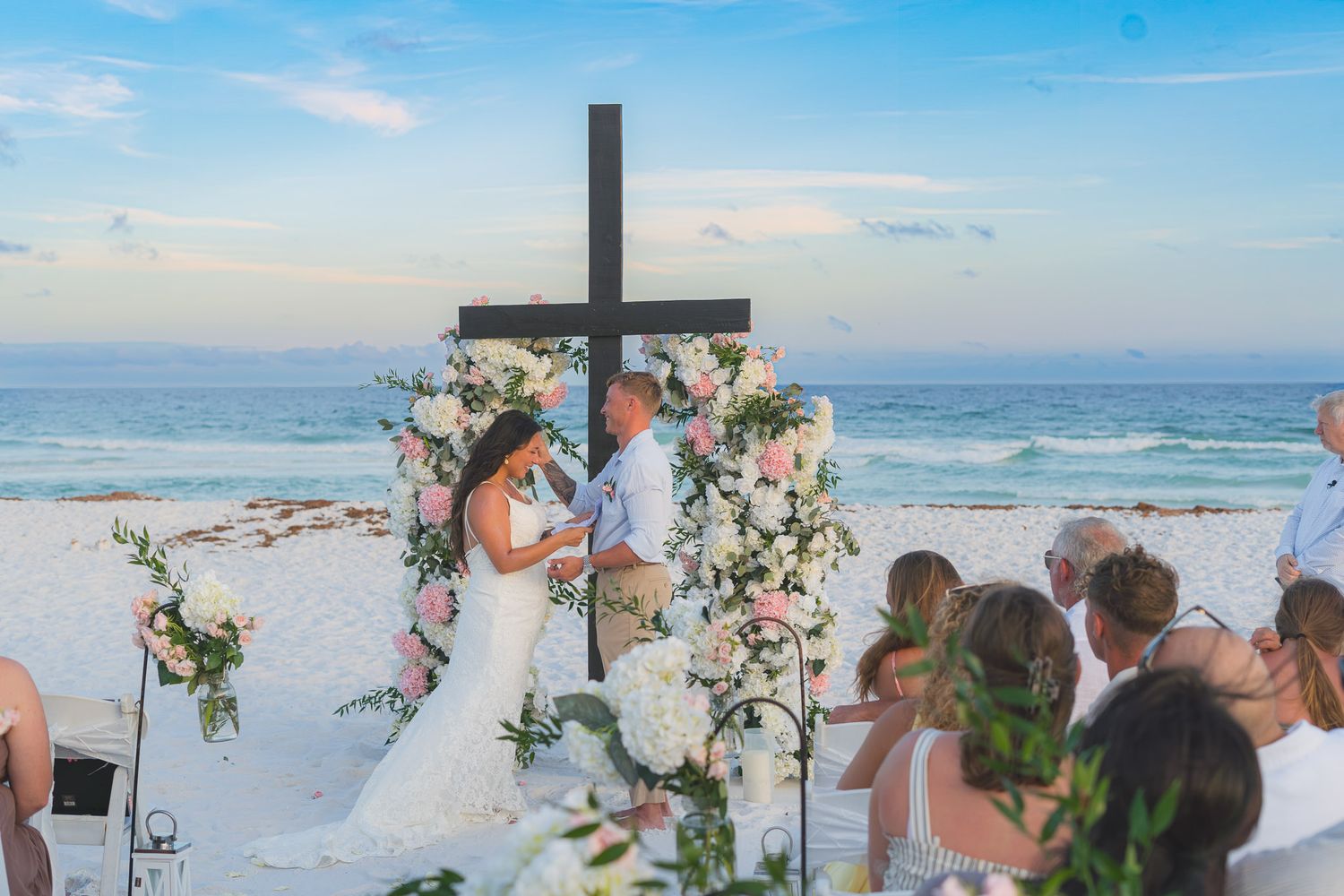 Couple exchanging vows at a beach wedding ceremony with floral arch and cross, guests watching.