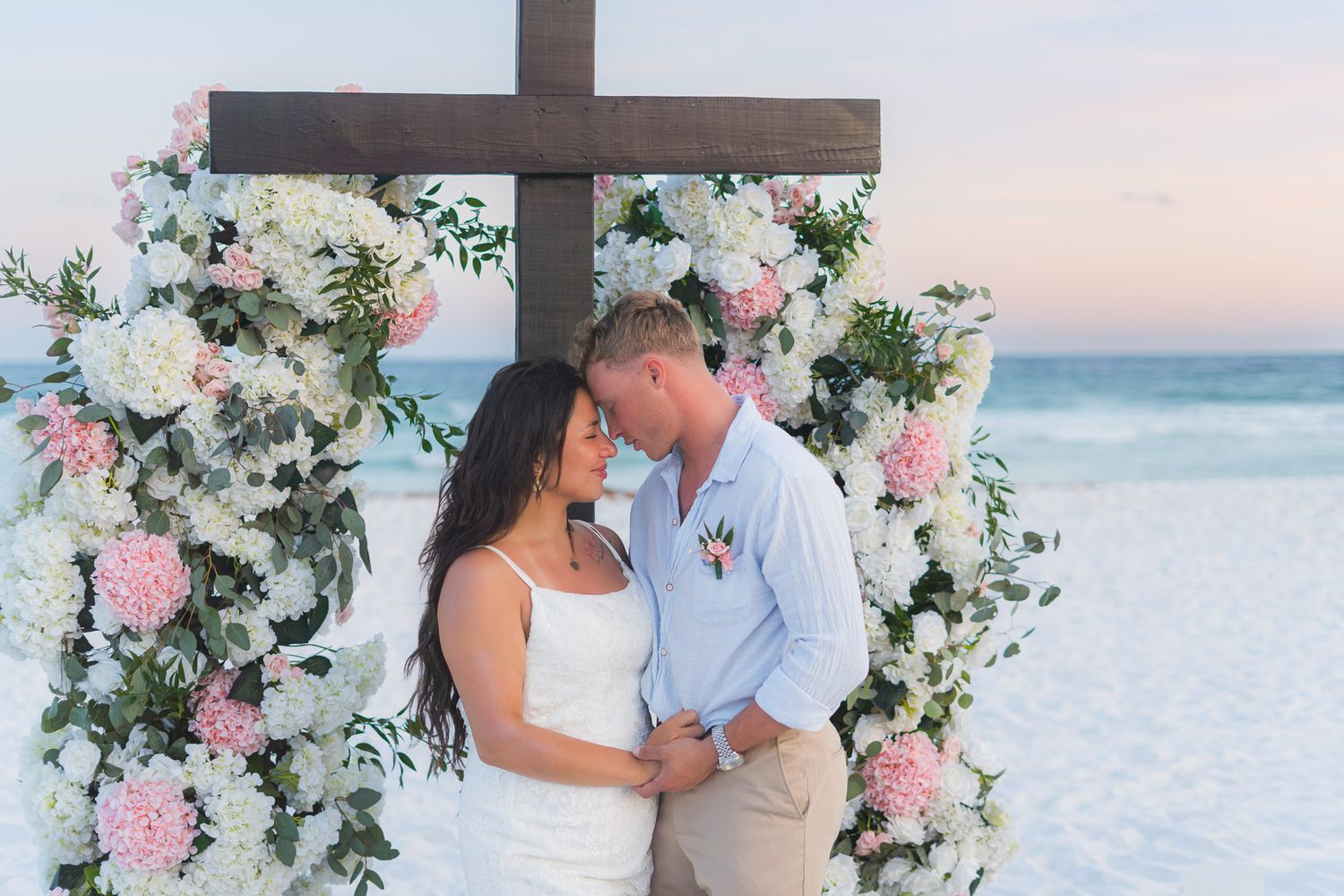 Couple sharing romantic moment at beach wedding ceremony under floral cross arch with pink and white flowers at sunset.