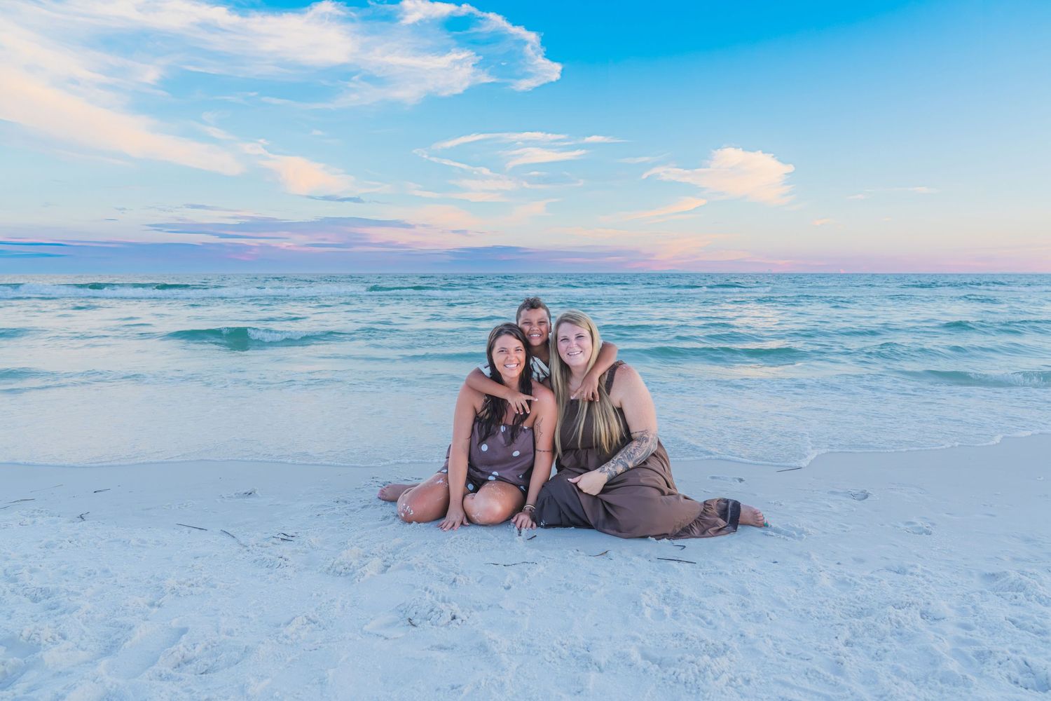 Three women smiling together on a white sandy beach at sunset with turquoise water and pastel sky.
