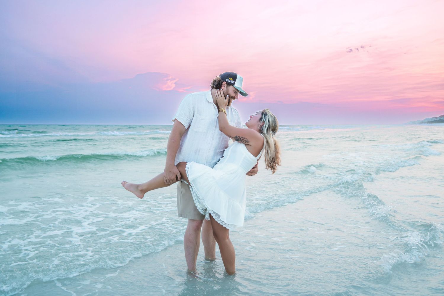 Couple sharing romantic kiss on beach at sunset, man lifting woman in white dress by ocean waves.