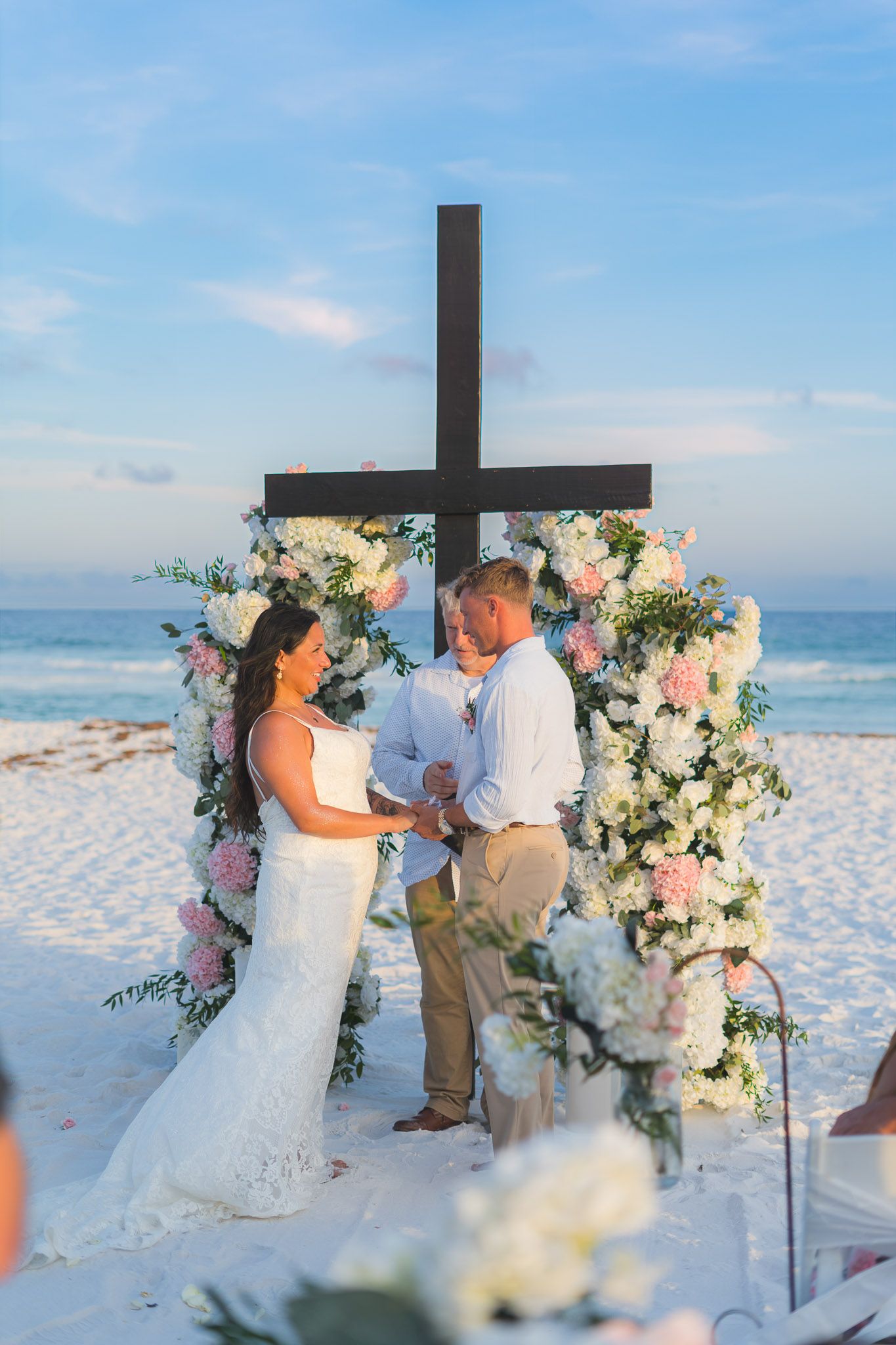 Bride and groom exchange vows at a beach wedding altar adorned with pink and white flowers and a wooden cross.