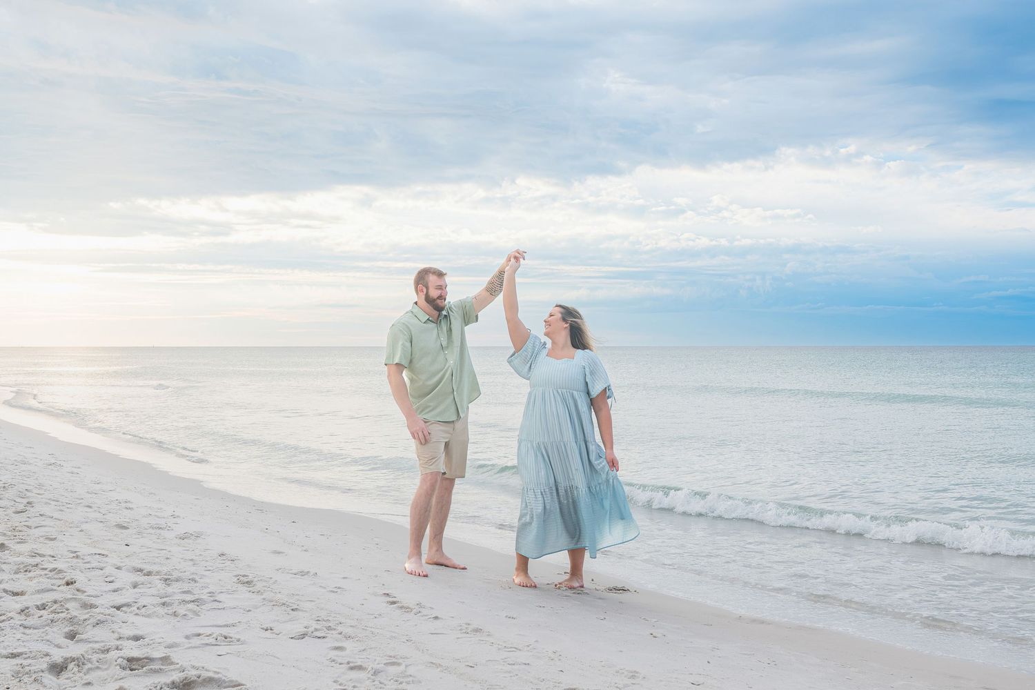 Couple dancing on a sandy beach with calm ocean waves and a soft blue sky backdrop.
