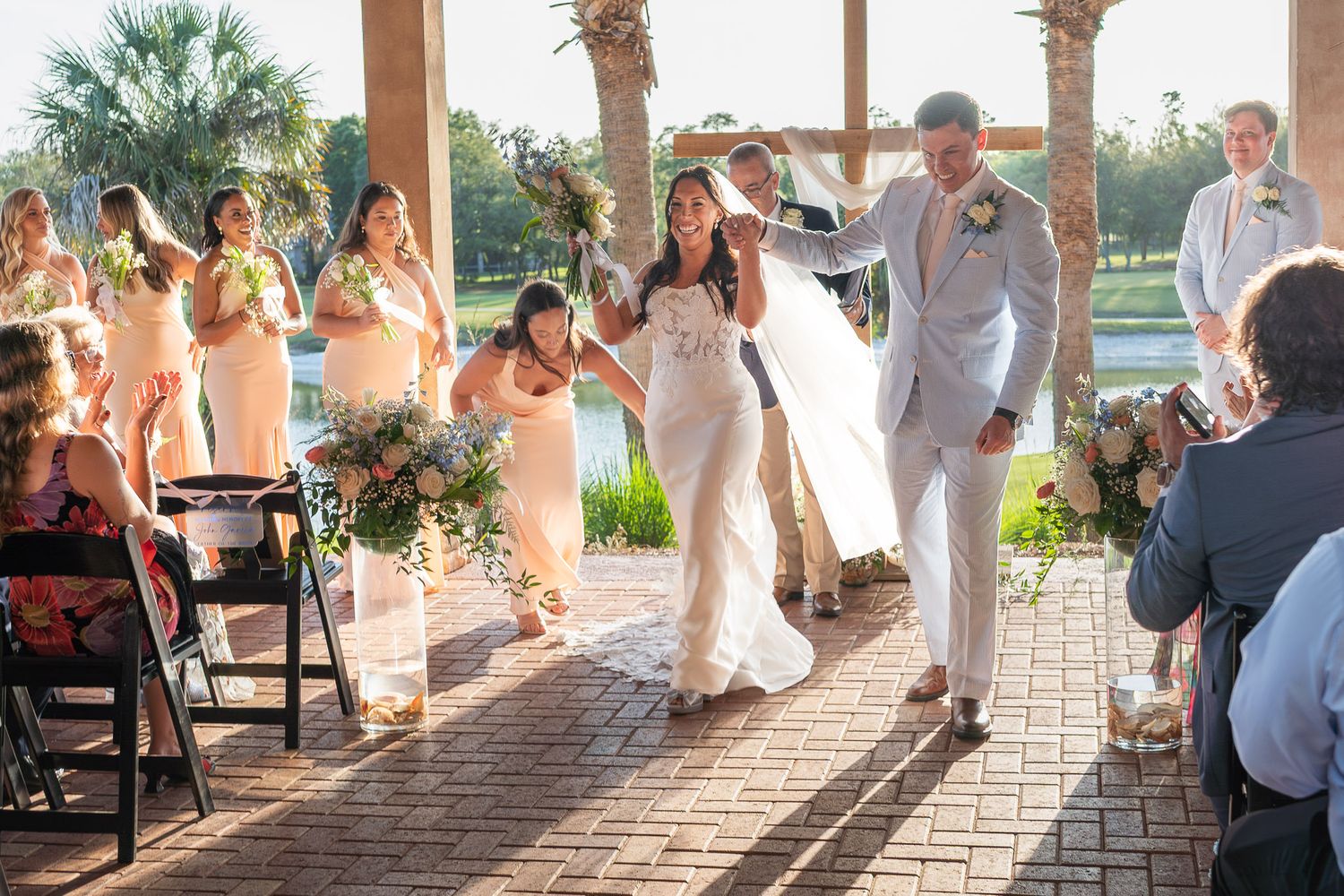 Newlyweds walk down aisle after wedding ceremony at outdoor venue with cheering guests.
