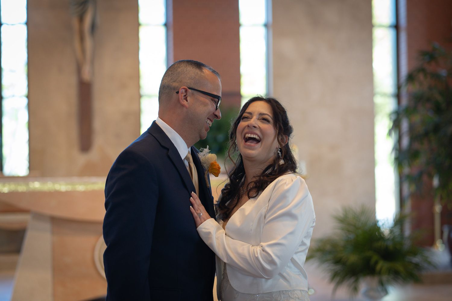 Bride laughs joyfully with groom during wedding portraits inside a sunlit church.
