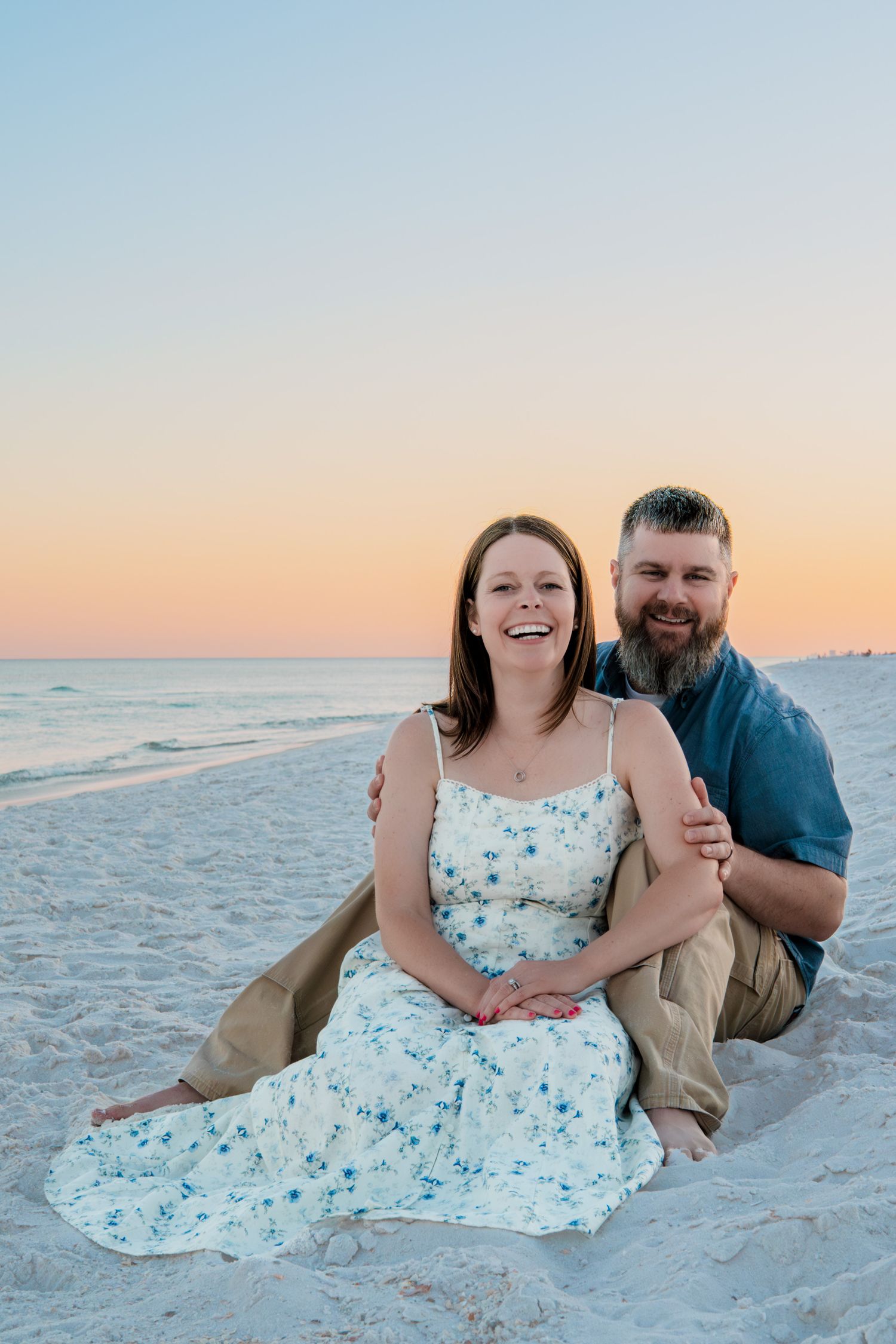 Couple sitting on white sand beach at sunset, woman in floral dress laughing, man with beard embracing her from behind.