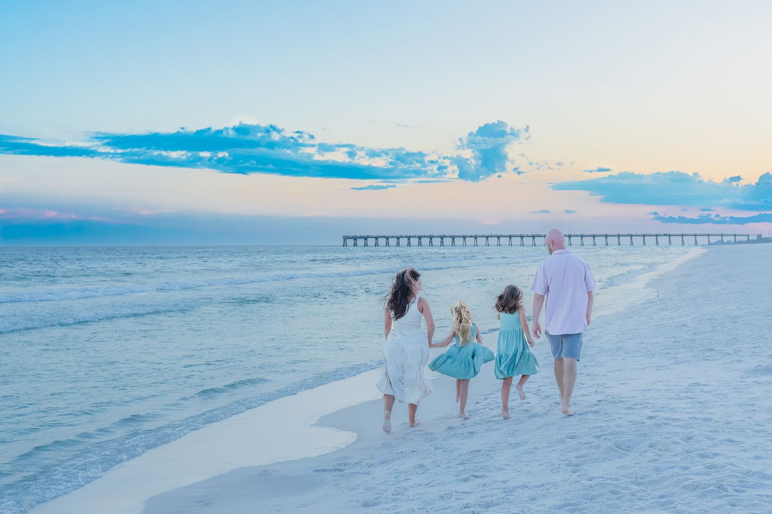 Family of four walking hand-in-hand on a white sandy beach at sunset with a pier in the background.
