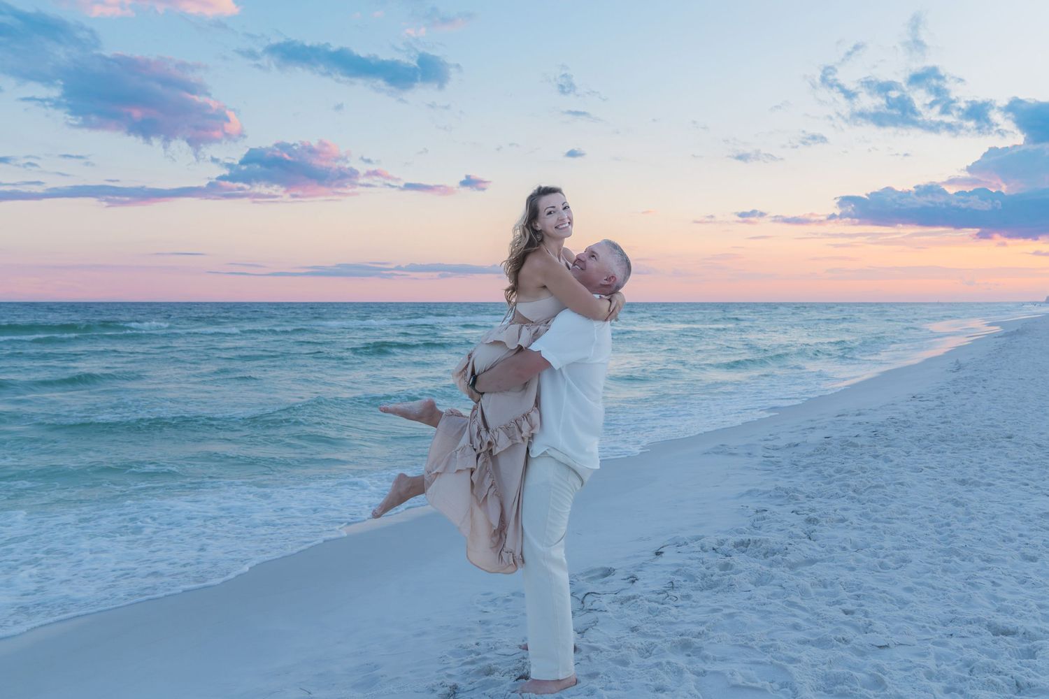 Couple embracing at sunset on a white sand beach with turquoise water and pastel pink sky.