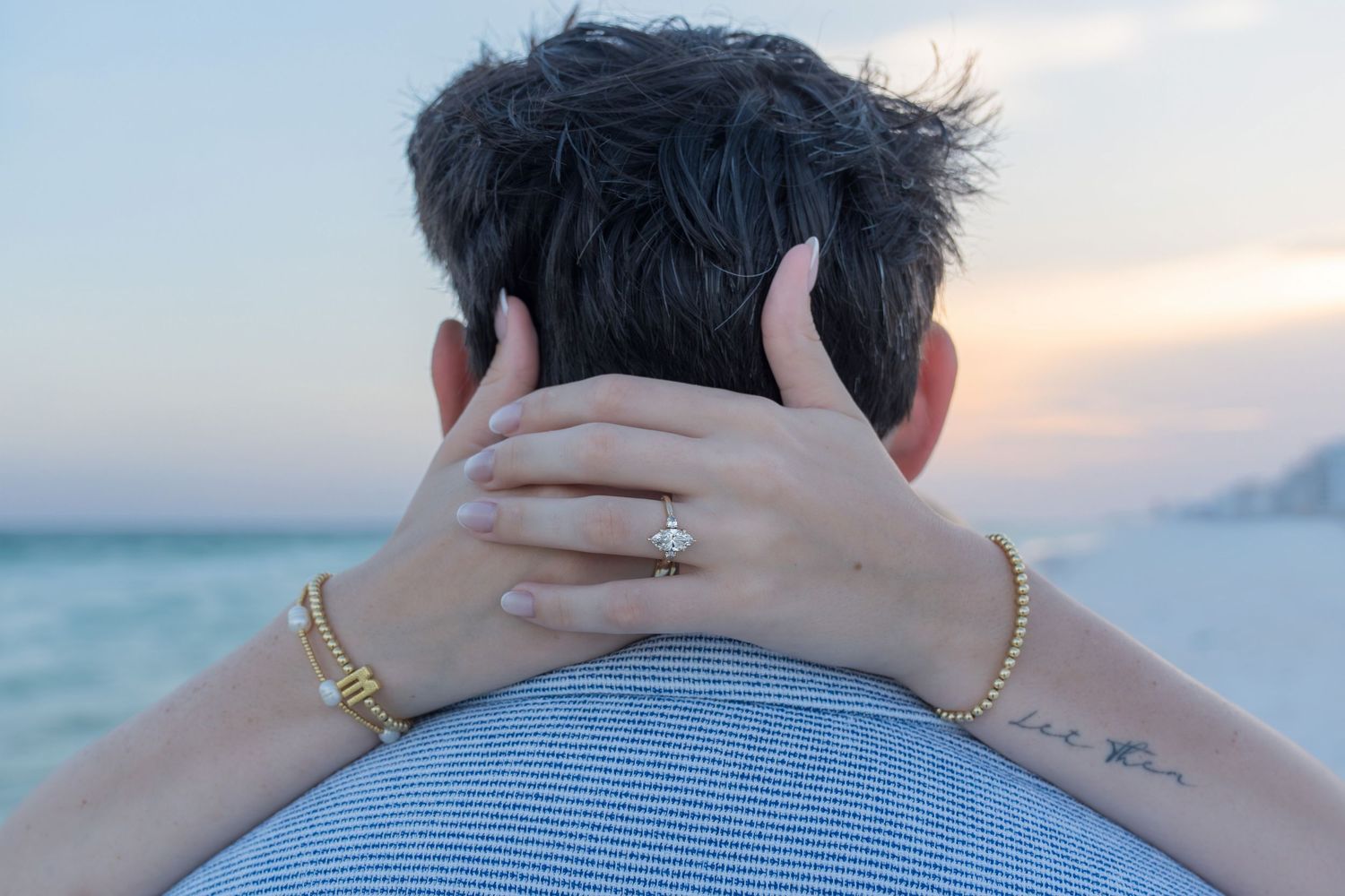 Couple embracing at sunset on a beach, woman showing diamond engagement ring and gold bracelet.