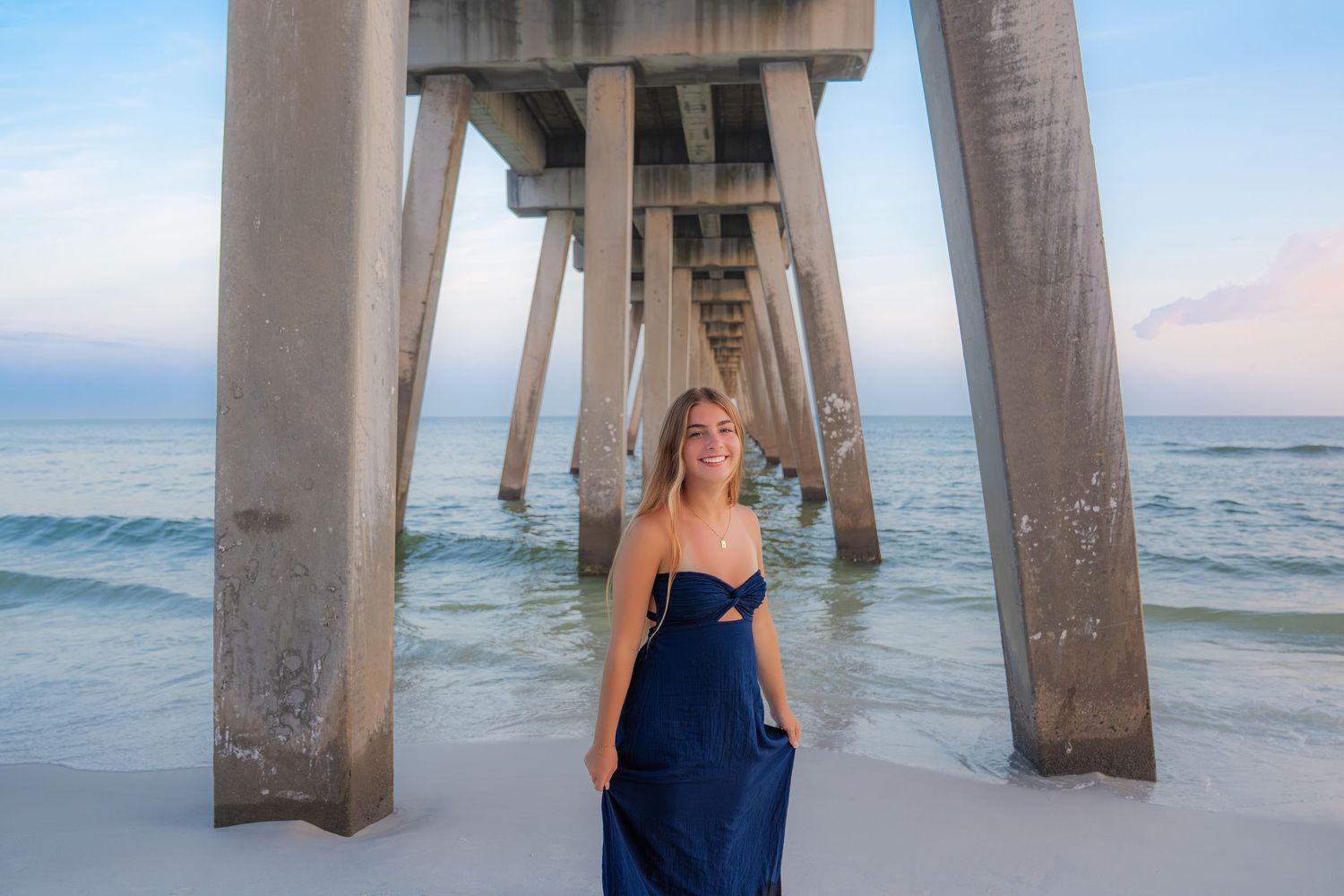 Woman in navy maxi dress smiling under a beach pier with ocean waves and pastel sky in background.