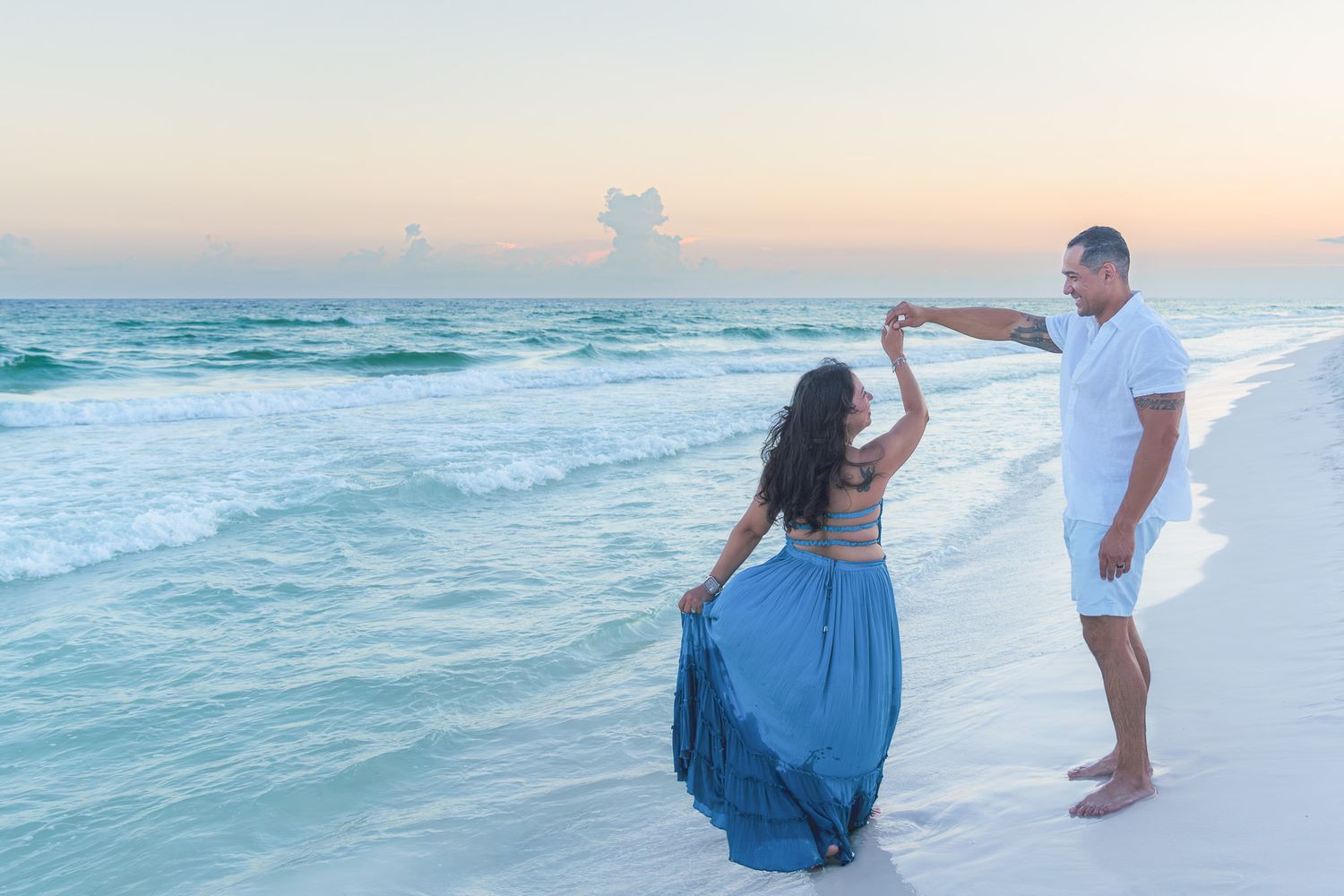 Couple dancing on a sandy beach at sunset with turquoise ocean waves in the background.