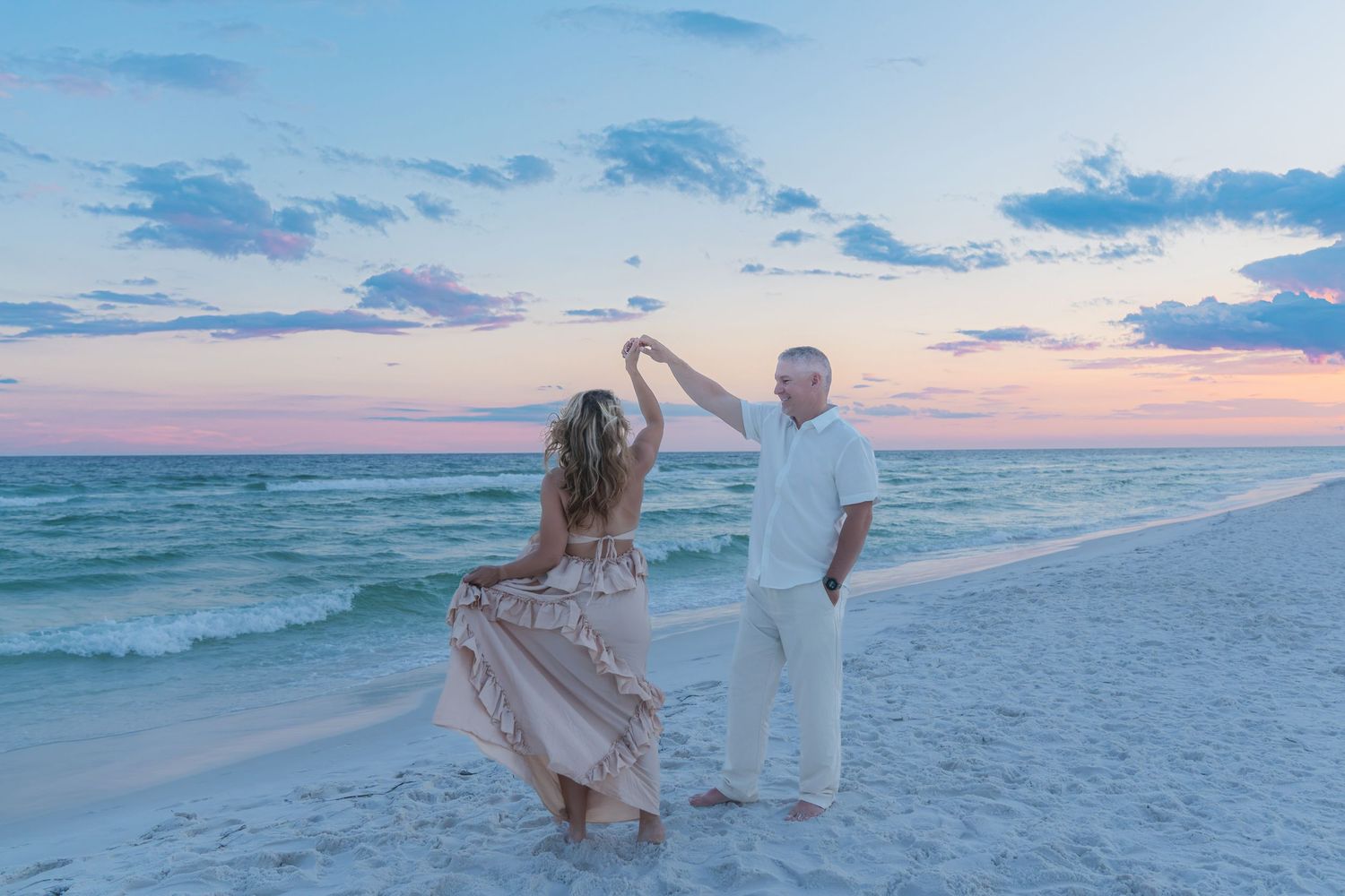 Couple dancing on a sandy beach at sunset with pastel skies and turquoise ocean waves in the background.