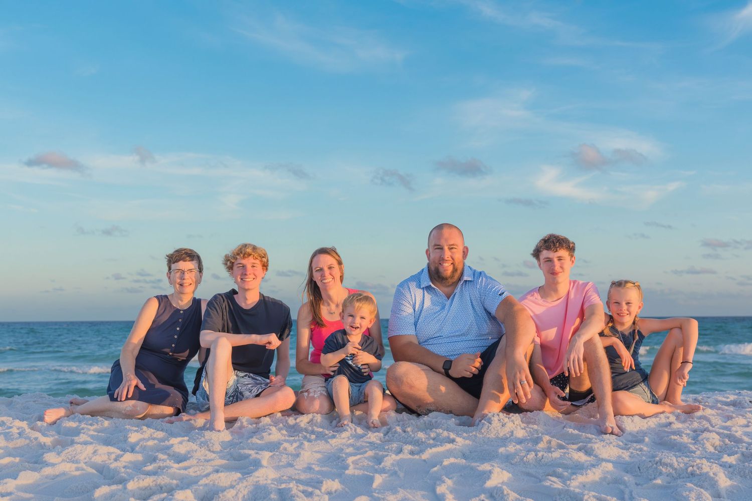 Family of seven posing together on white sandy beach at sunset with turquoise ocean waves in background.