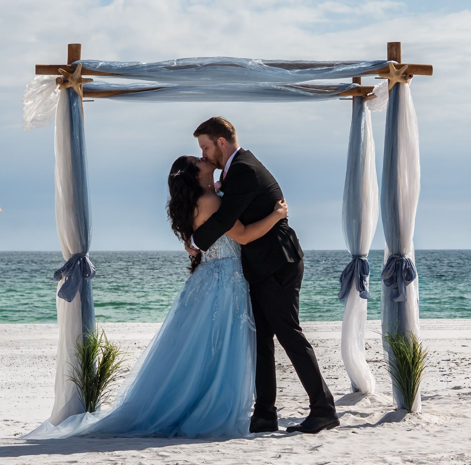 Bride and groom share a kiss under a rustic wooden arch decorated with blue ribbons on a sunny beach.