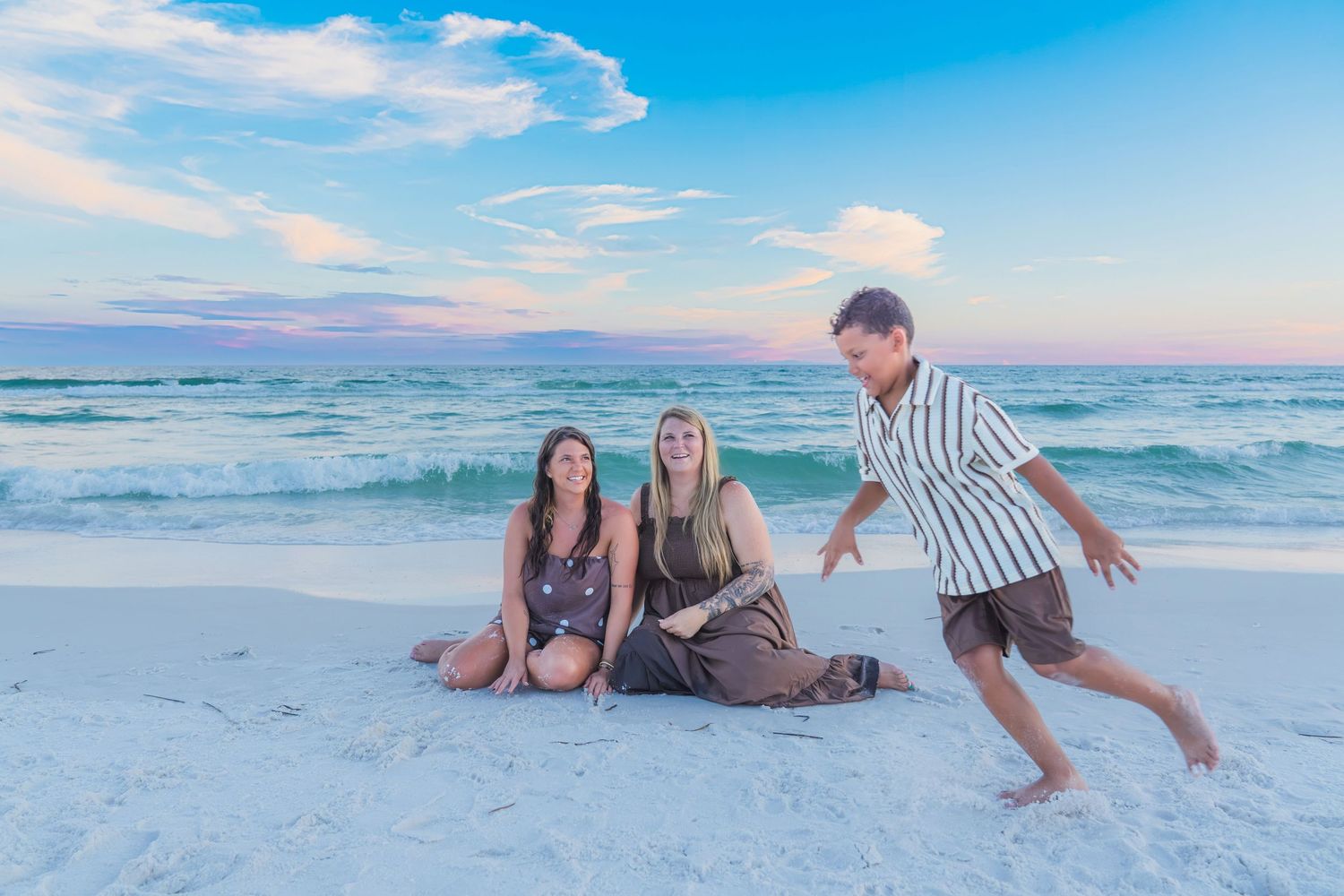 Two women sit on white sand beach at sunset while a man playfully runs toward them with ocean waves behind.