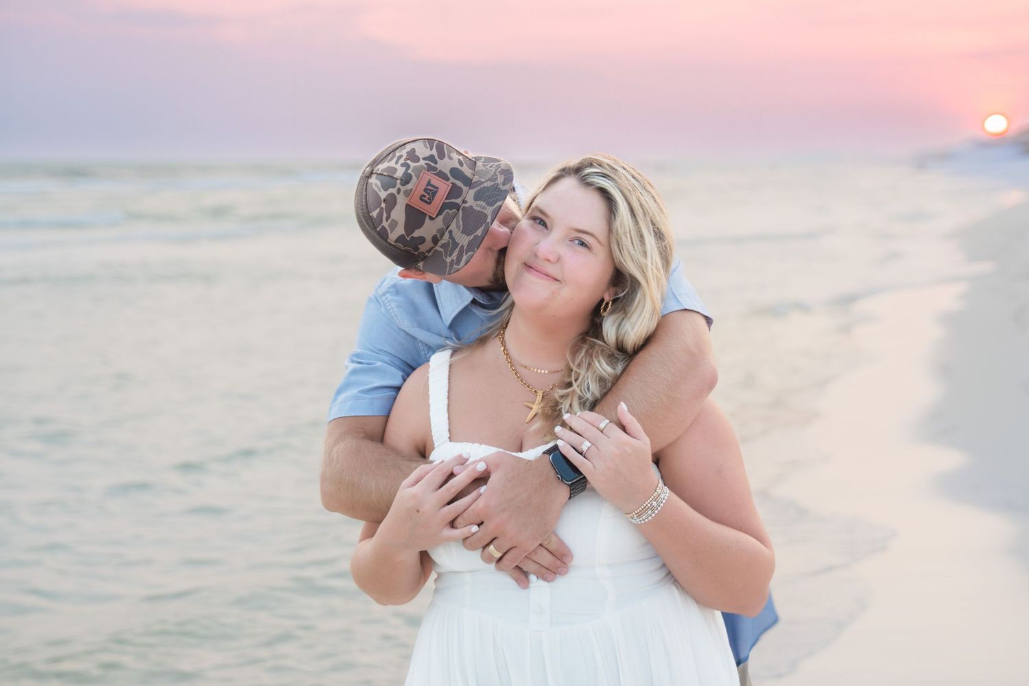 Couple embracing on a beach at sunset, woman in white dress smiling as man kisses her cheek.