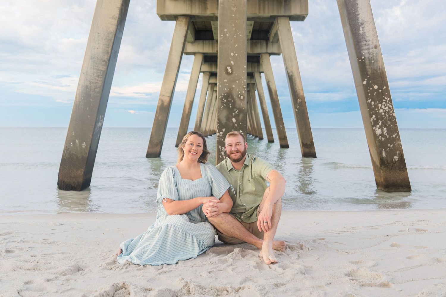 Couple sitting together under a beach pier on white sand with calm ocean waters and cloudy sky backdrop.