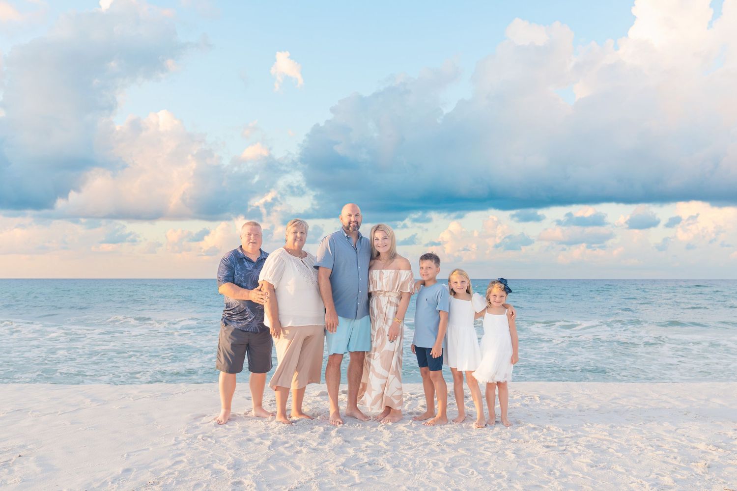 Family of seven posing together on a white sand beach with calm turquoise water and pastel cloudy sky.