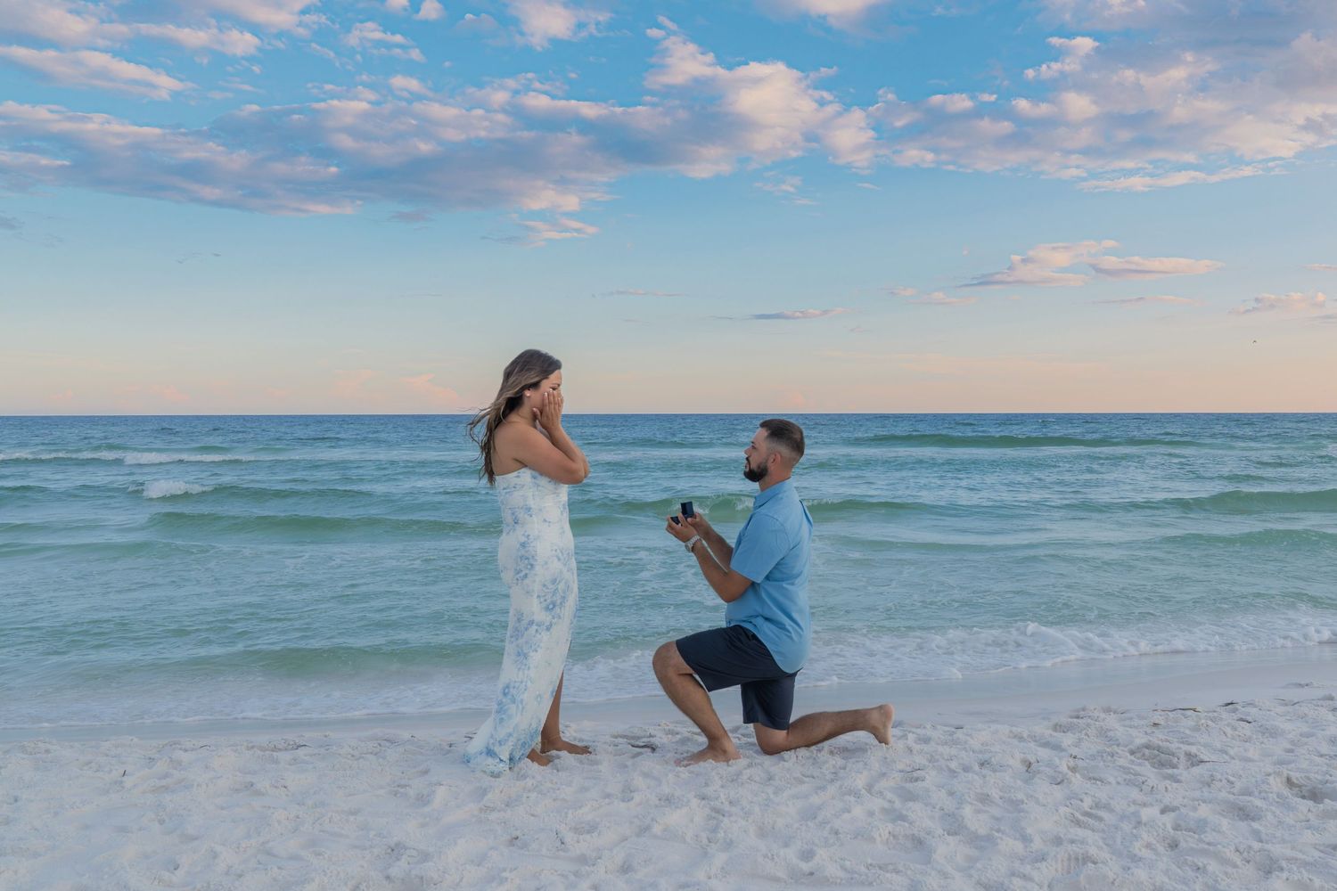 Man proposing on one knee to woman on a sandy beach at sunset with ocean in background.
