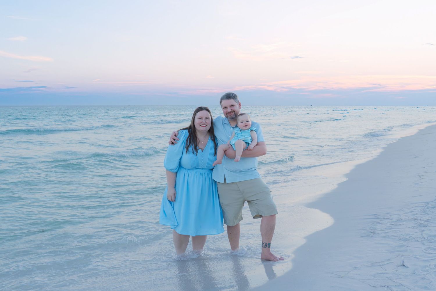 A couple stands on a sandy beach at sunset, the woman in a blue dress and man holding a baby, with pastel skies and calm ocean waves.