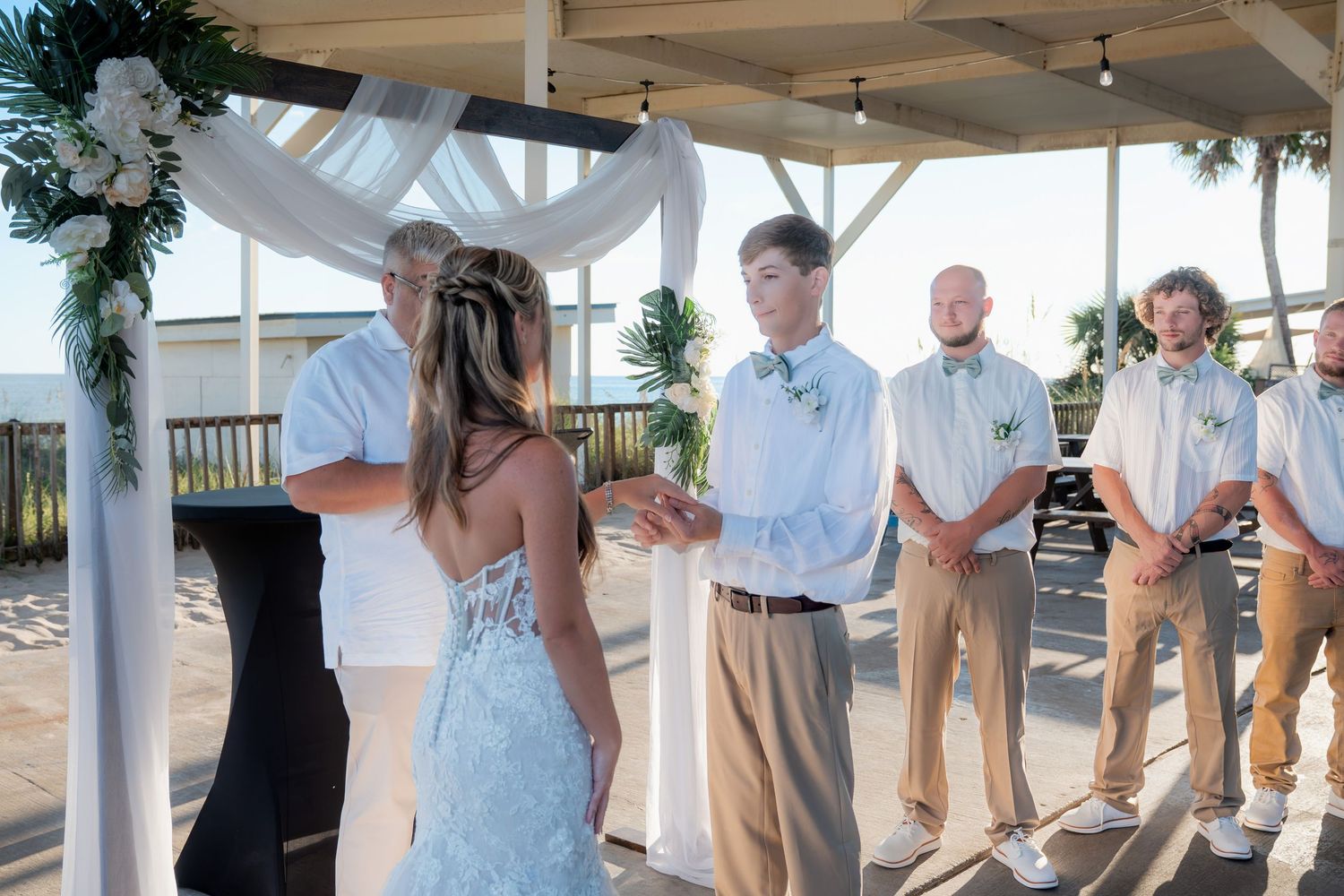 A bride and groom exchange vows at an outdoor beach wedding ceremony with groomsmen standing nearby.
