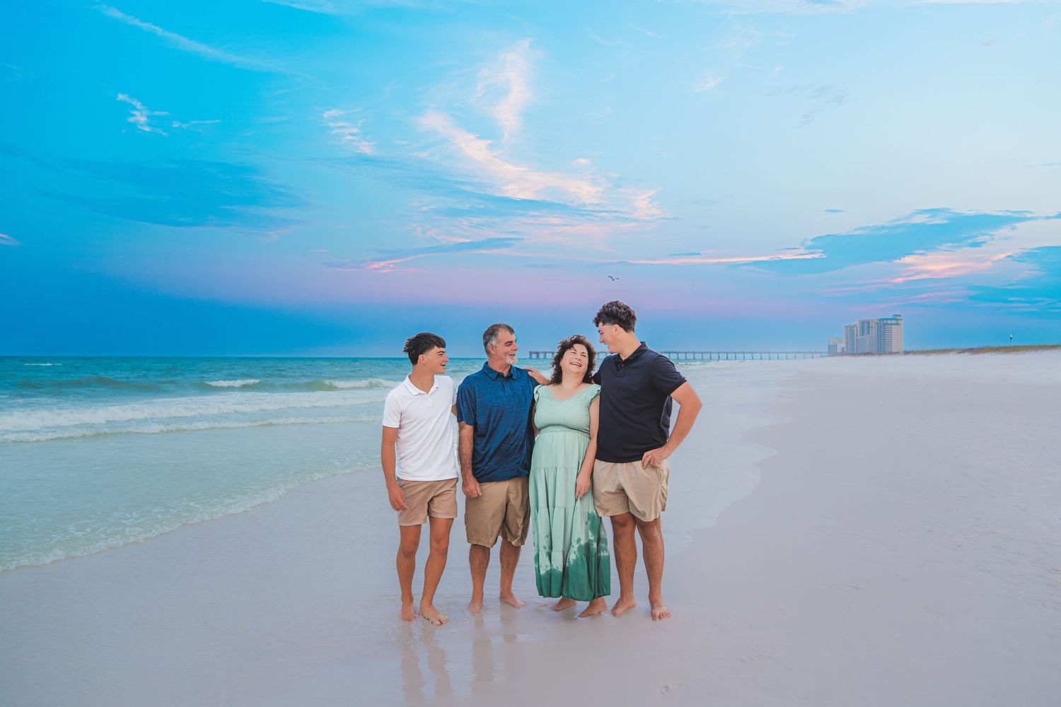 Family of four posing together on a white sand beach at sunset with pastel pink and blue skies.