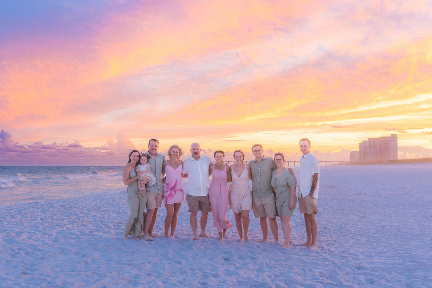 Large family group posing on a sandy beach during a vibrant pink and orange sunset near the ocean.