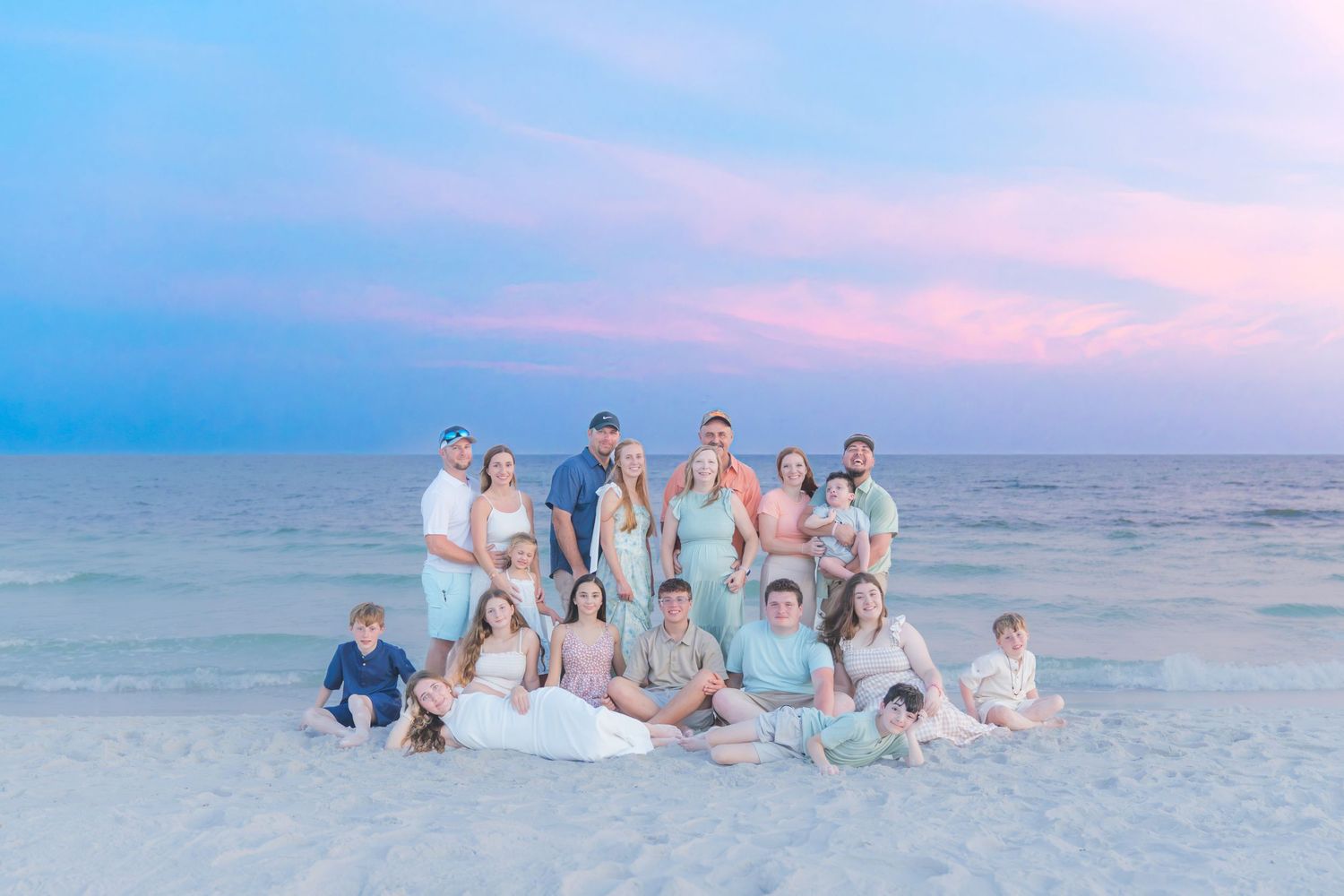 Large family posing together on a white sand beach at sunset with pink and blue pastel sky over calm ocean waters.