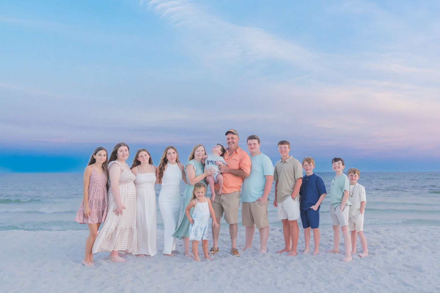 Large family posing together on a white sand beach at sunset with pastel pink and blue sky over calm ocean waters.