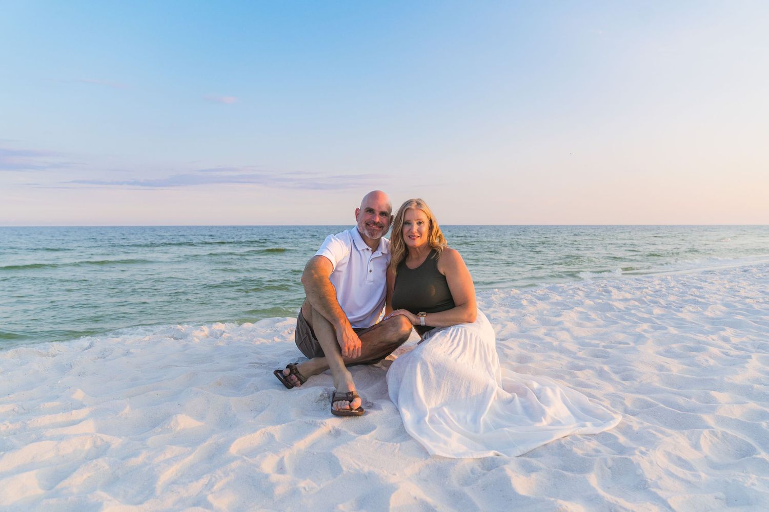 Couple sitting together on white sandy beach at sunset with turquoise ocean waves in the background.