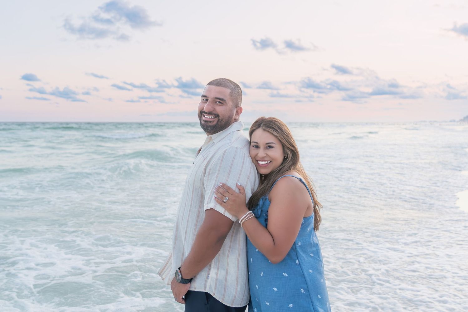 Smiling couple posing back-to-back on a sandy beach with ocean waves and pastel sunset sky behind them.