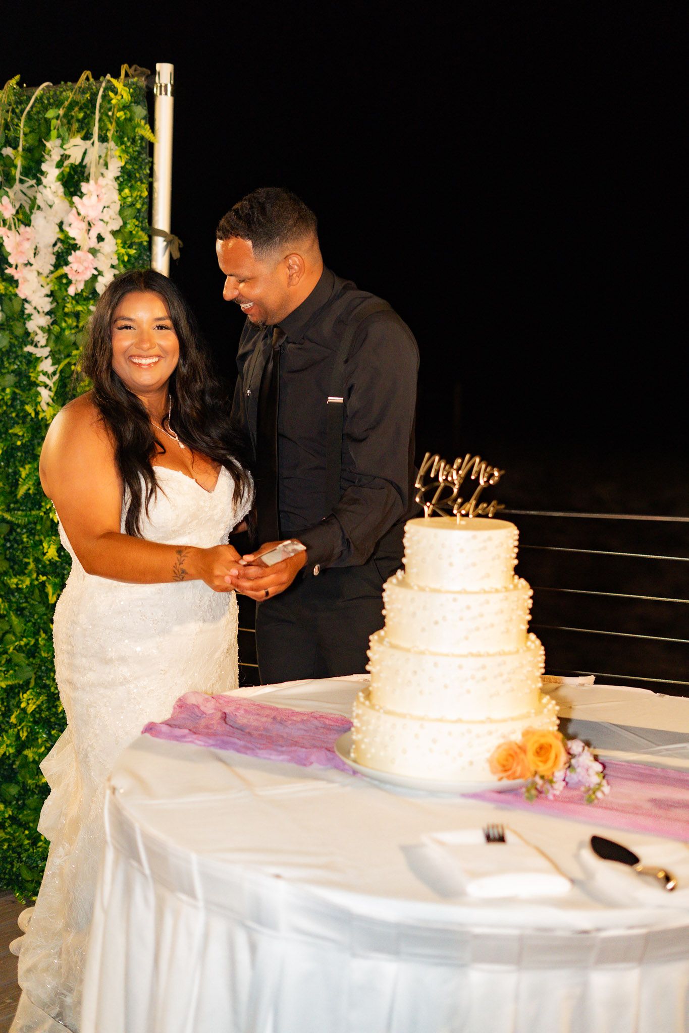 Smiling bride and groom prepare to cut their elegant four-tier white wedding cake at their reception.