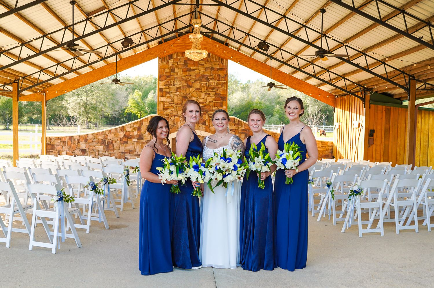 Bride with four bridesmaids in navy blue gowns holding white bouquets inside rustic wedding venue pavilion.
