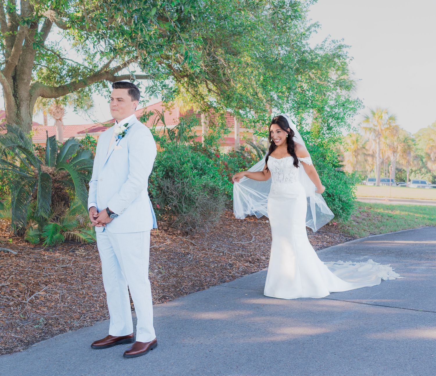 A bride in a white gown and groom in a white suit pose for a first look photo on a tree-lined path.