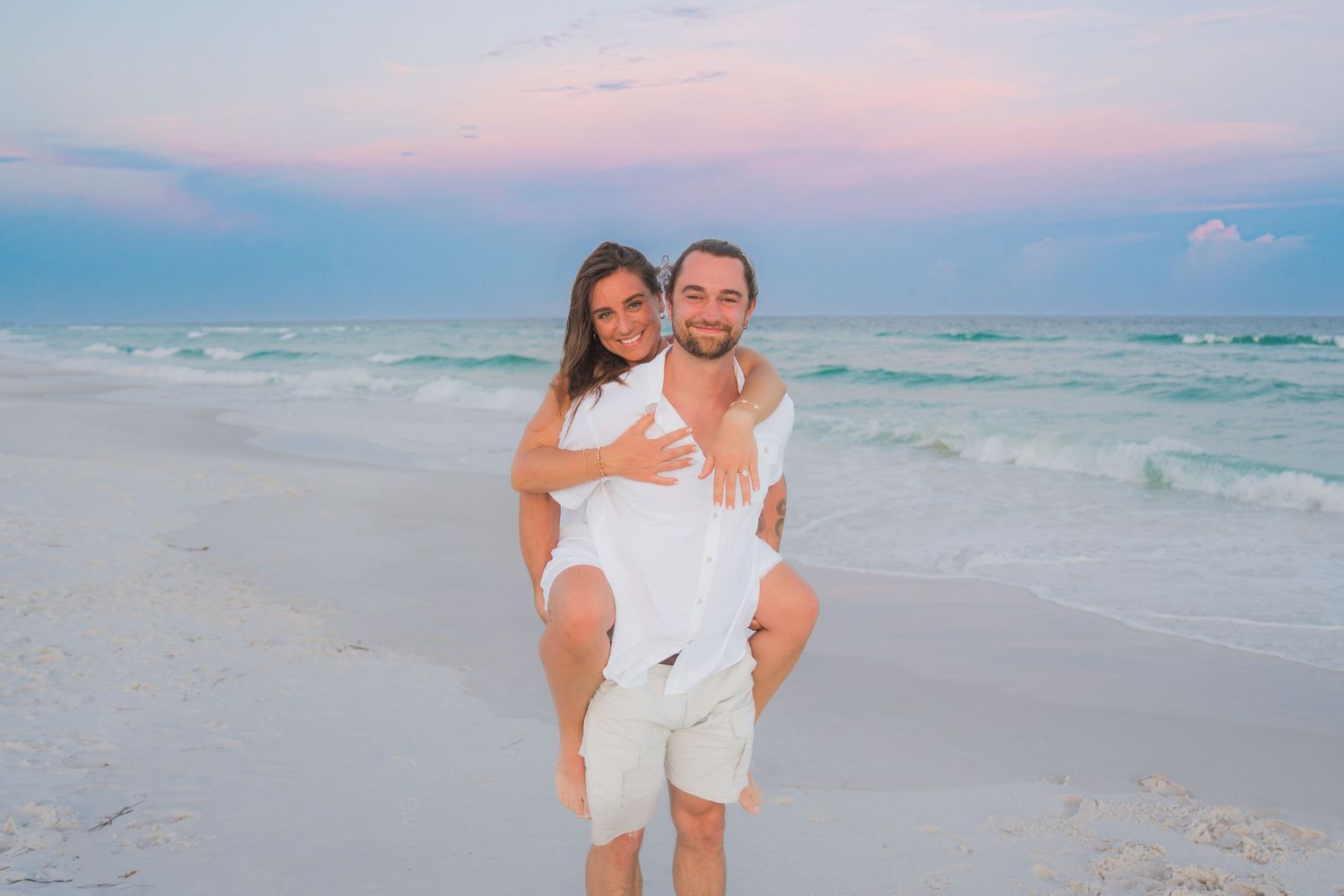 Couple in white outfits on a sandy beach at sunset, man giving woman a piggyback ride with pastel sky.