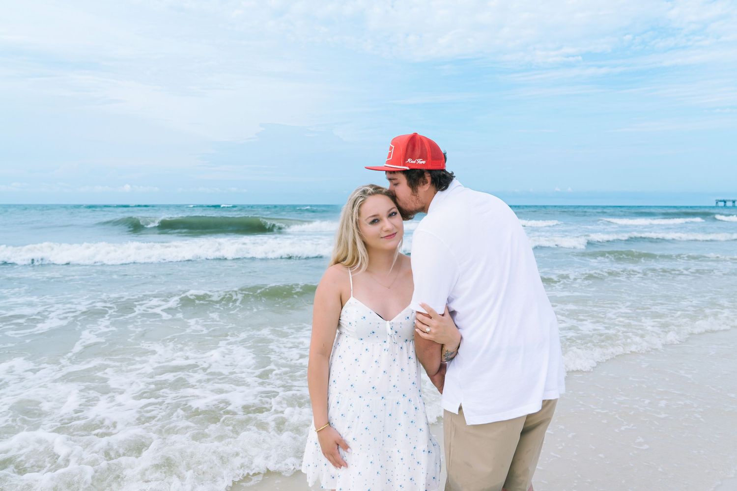 Couple embracing on a sandy beach, woman in white sundress, man in red cap and white shirt, ocean waves behind them.