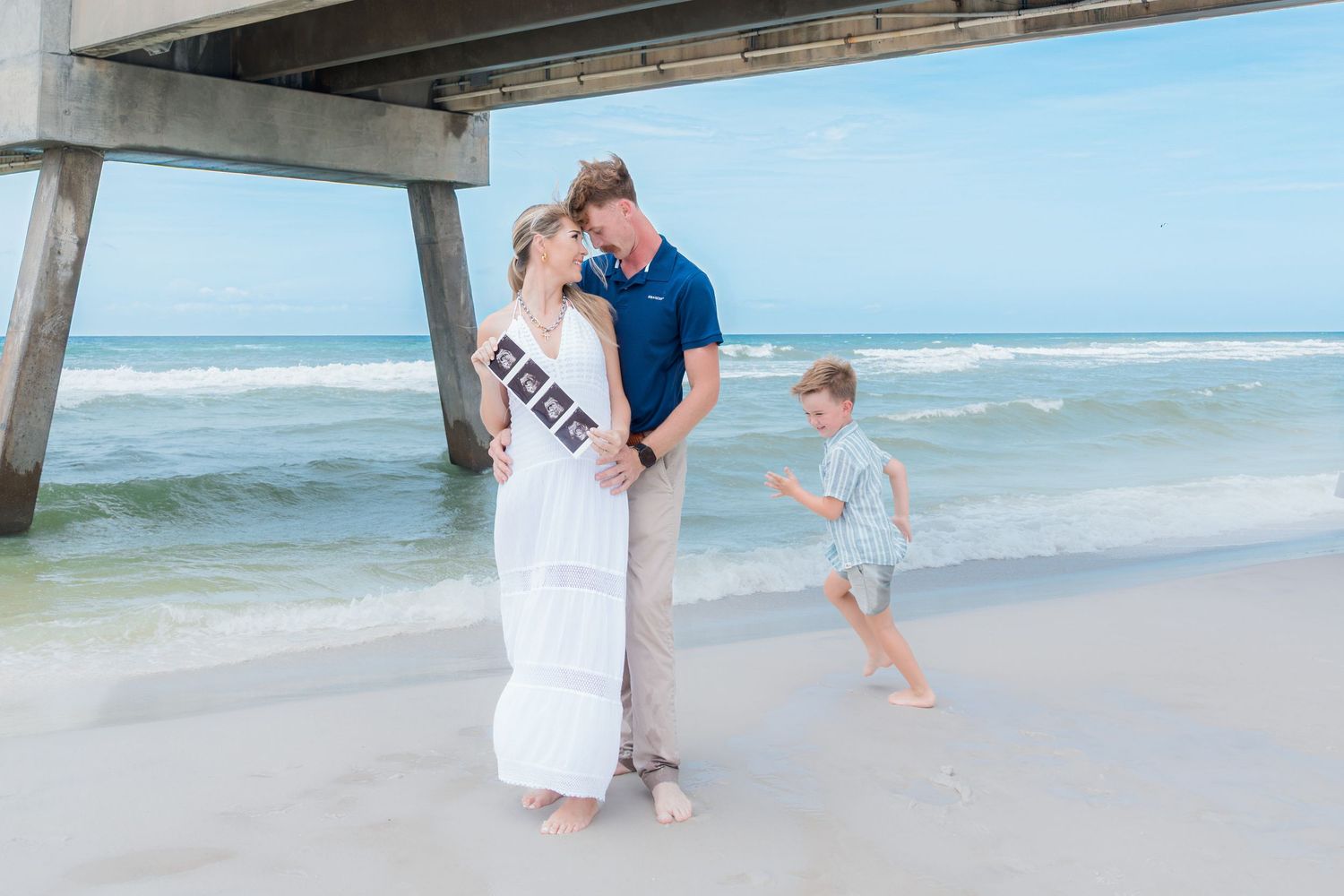 Pregnant woman holding ultrasound photos with partner and child on sunny beach near pier.