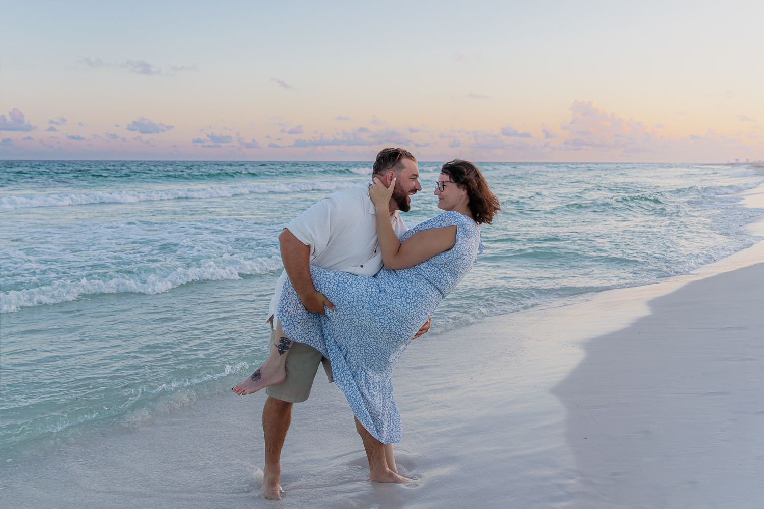 Couple sharing a romantic moment on a white sand beach at sunset, man lifting woman in a blue floral dress.