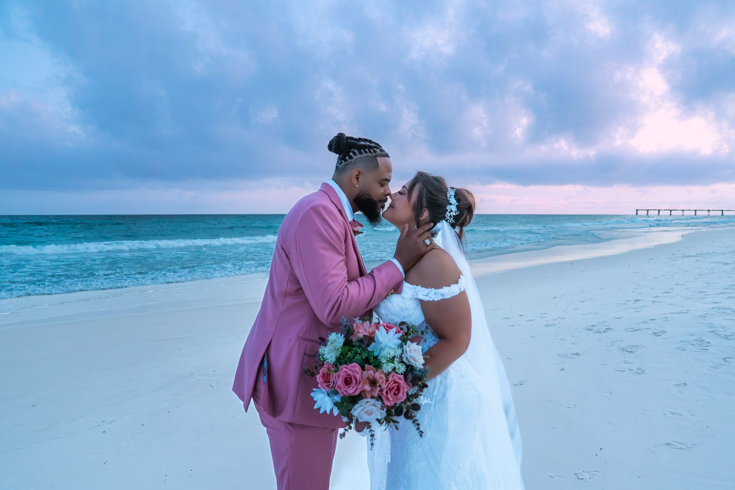 Newlywed couple kissing on a white sand beach at sunset, groom in pink suit, bride in white gown holding floral bouquet.