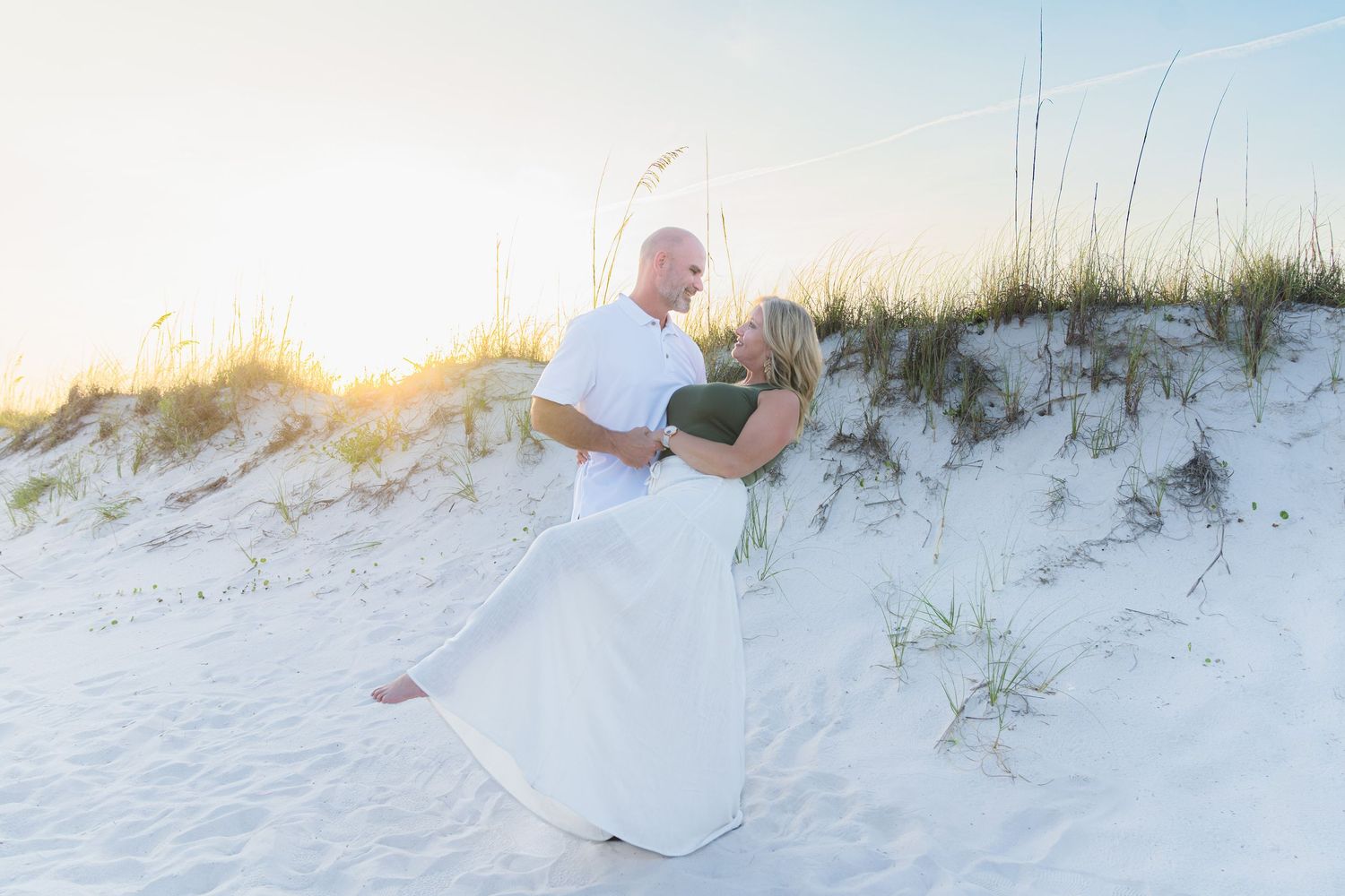 Couple in white attire embracing on sandy beach dunes at golden sunset with sea oats in background.