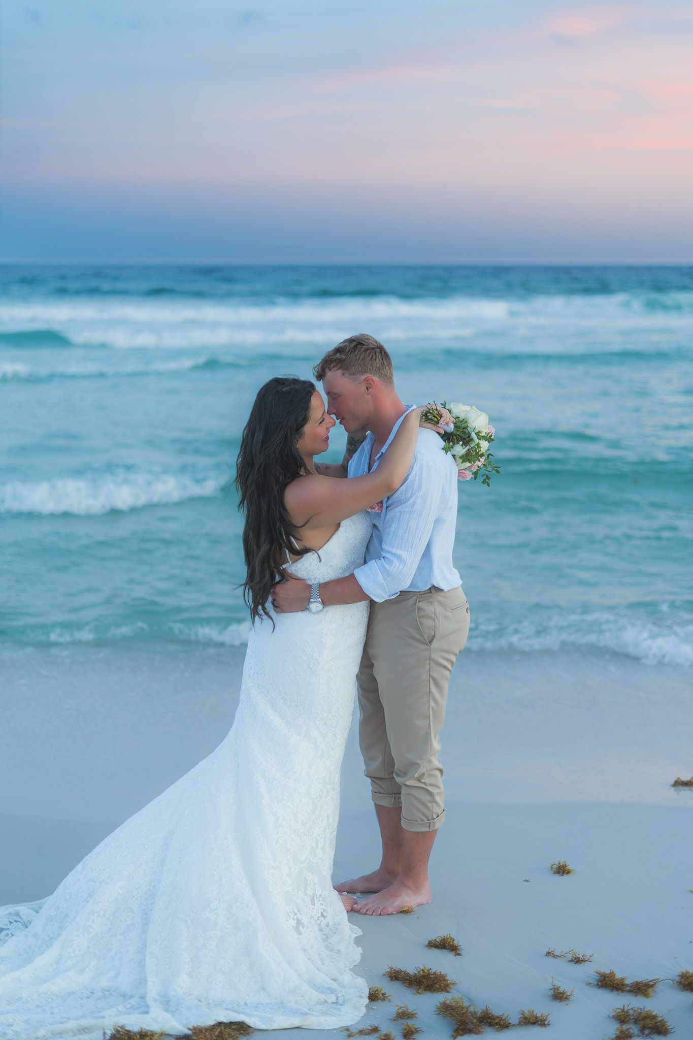 Bride in white lace gown and groom in khakis share romantic moment on sandy beach at sunset.