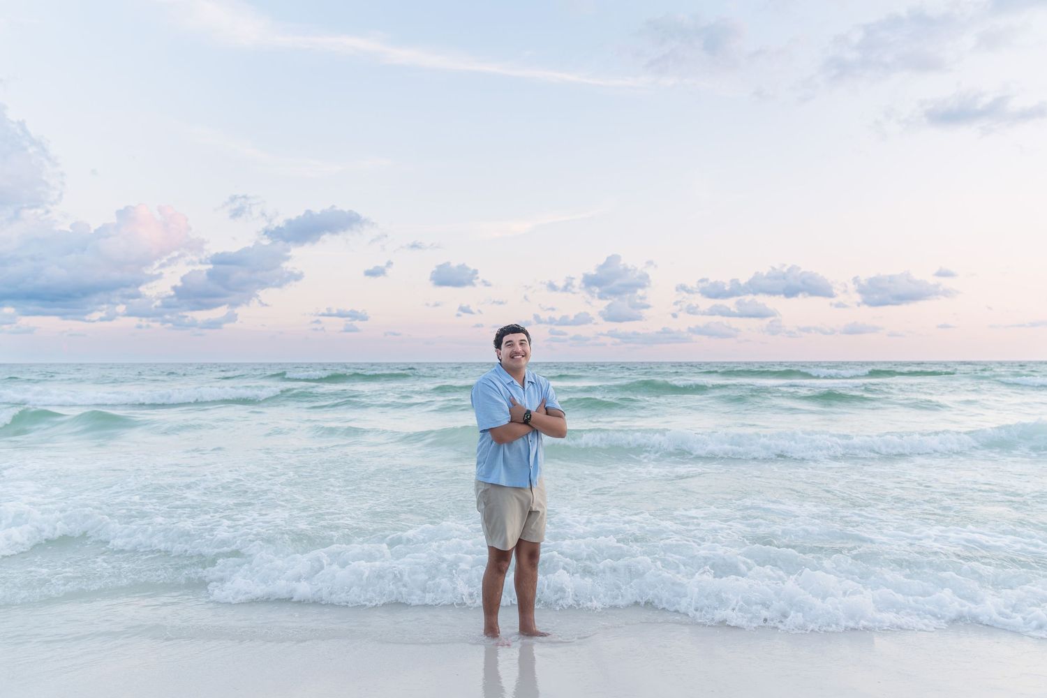 Man standing on a sandy beach with arms crossed, wearing a light blue shirt and khaki shorts, with ocean waves behind him.