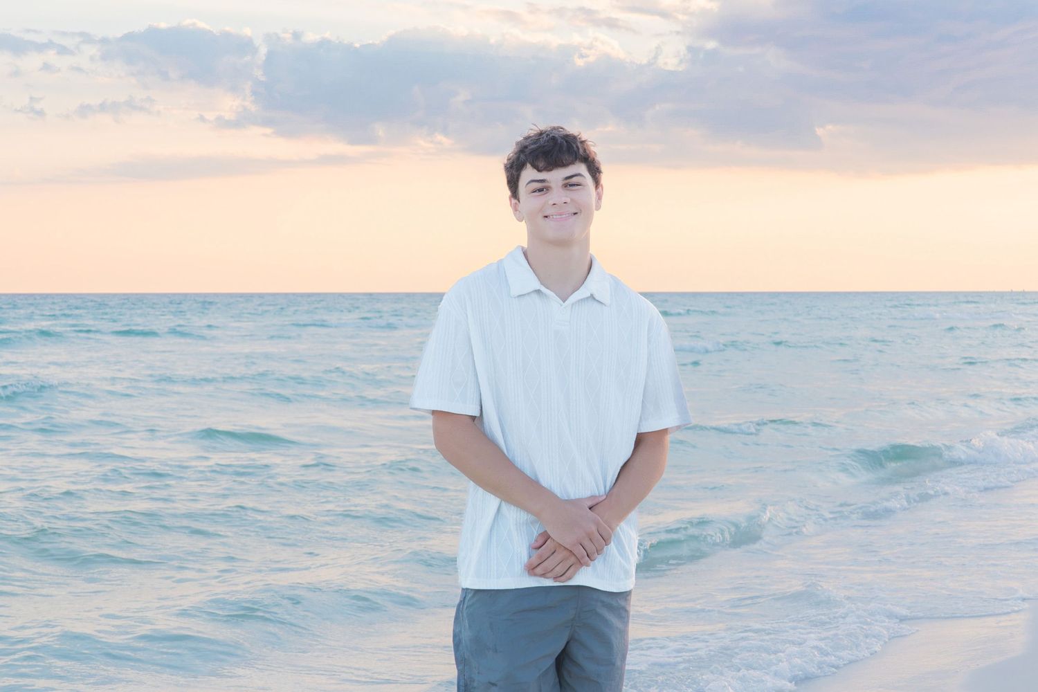 Smiling teenage boy in white polo shirt standing on a sandy beach at sunset with calm ocean waves behind him.