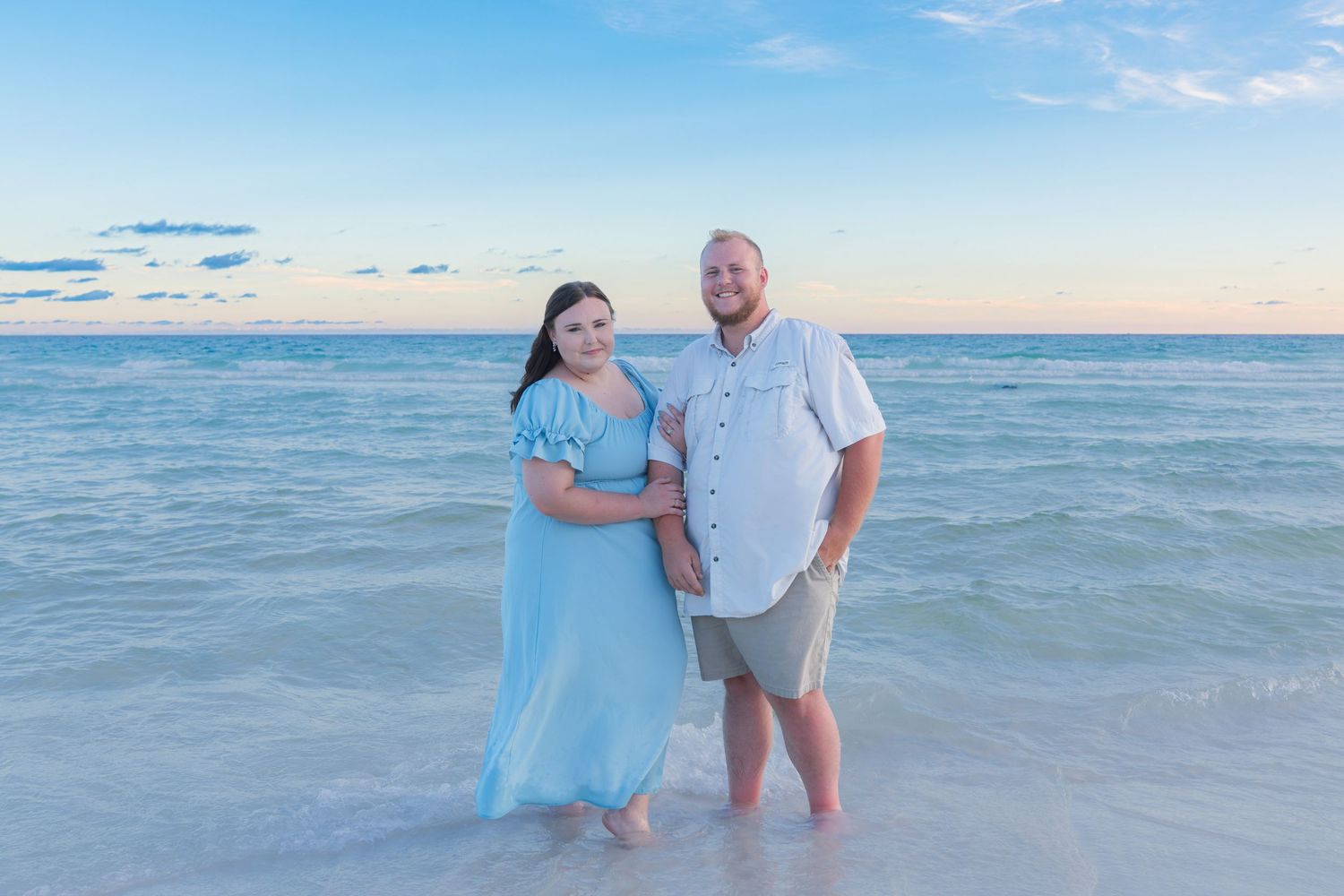 Couple standing in shallow ocean water at sunset, woman in blue dress and man in white shirt, beach photography.