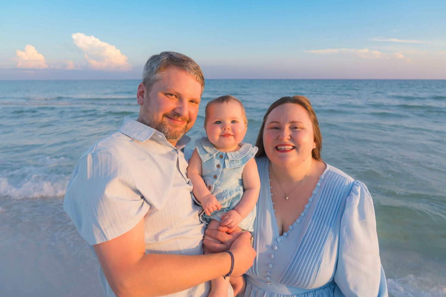 Family of three smiling on a beach at sunset, parents holding baby in blue outfit with turquoise ocean waves behind them.