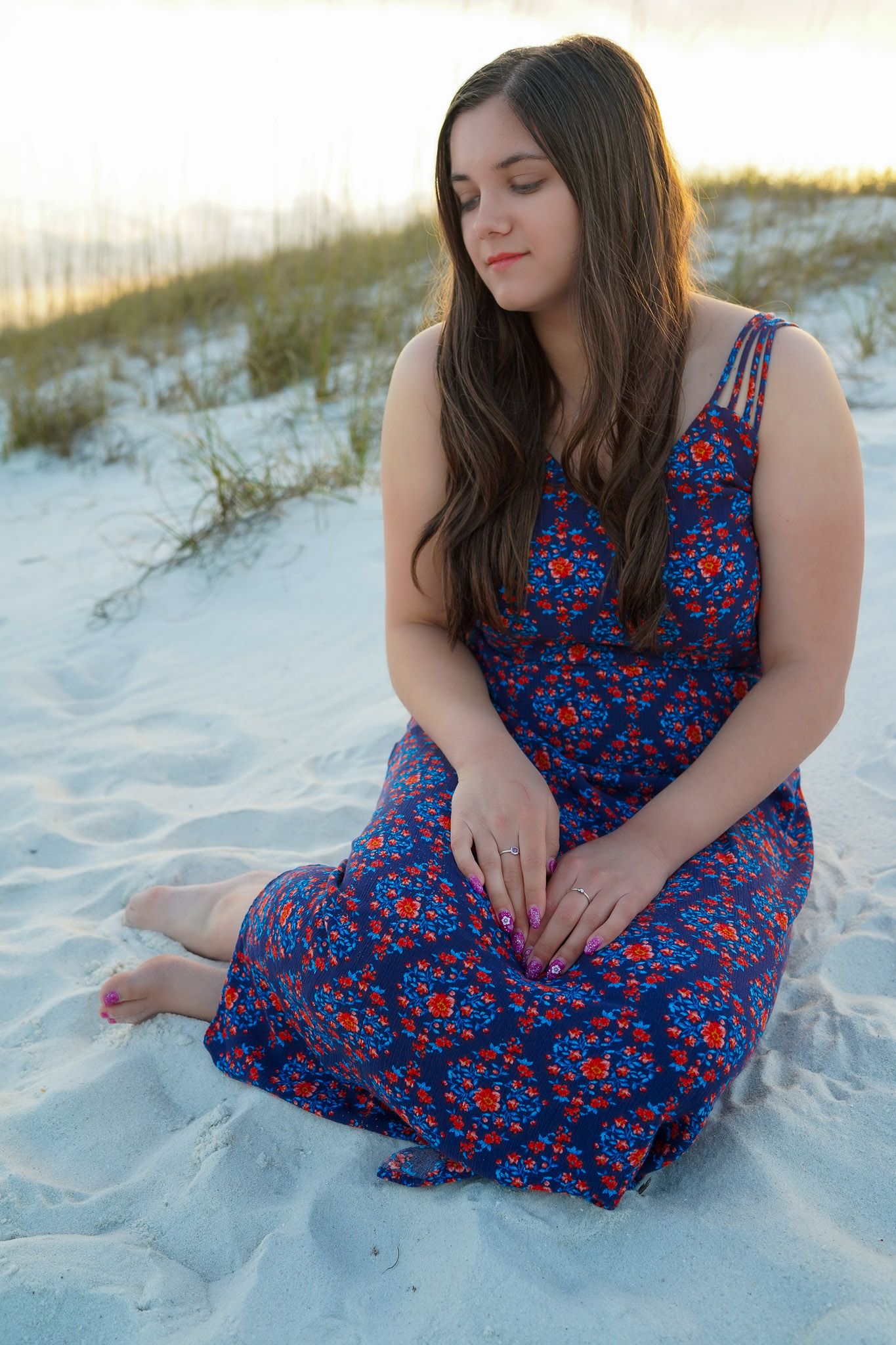Young woman in blue floral maxi dress sitting on white sand beach at sunset.