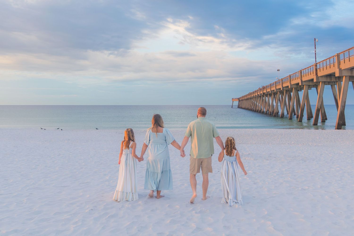Family of four holding hands on white sand beach near a pier at sunset in pastel outfits.