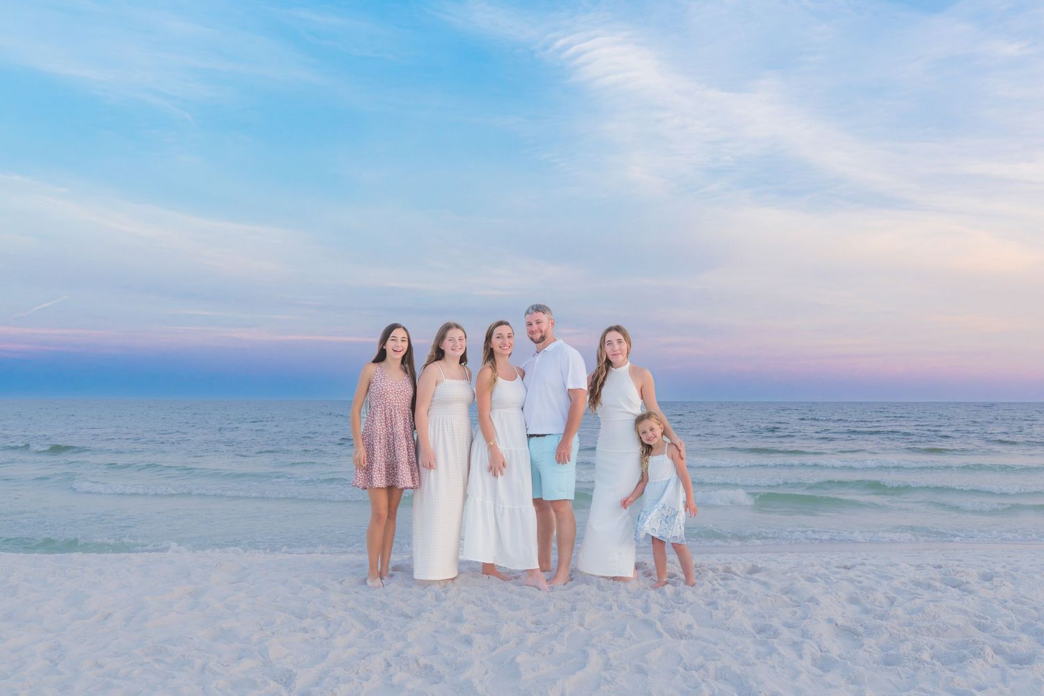 Family of six posing on a white sand beach at sunset with pastel pink and blue sky over calm ocean waters.