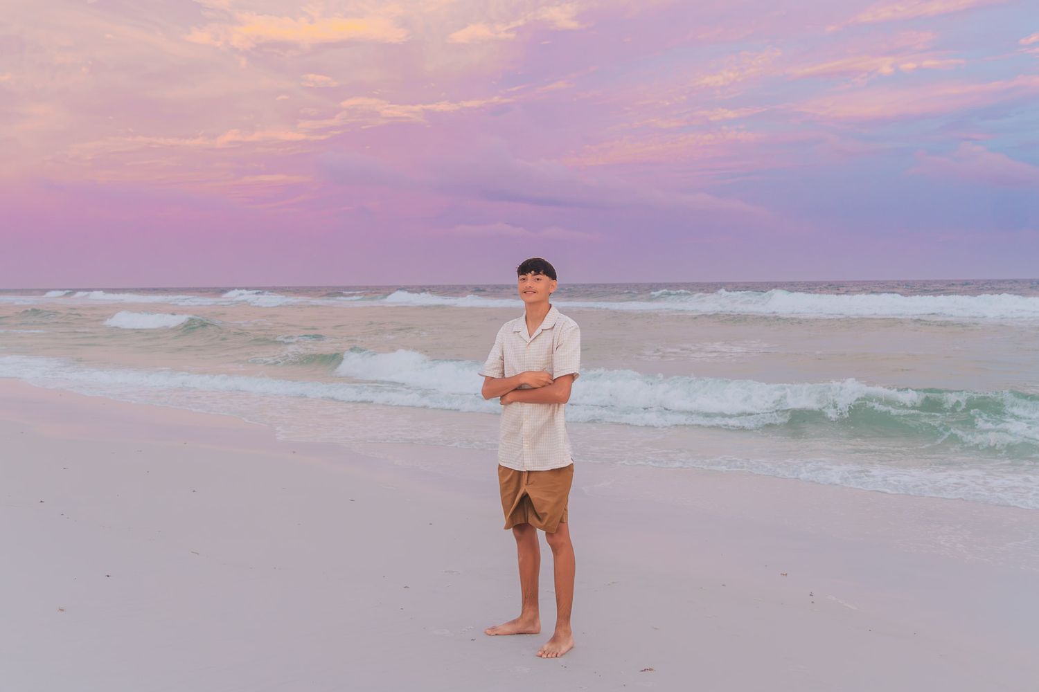 Man standing on white sand beach at sunset with pink and purple sky, ocean waves, wearing white shirt and brown shorts.