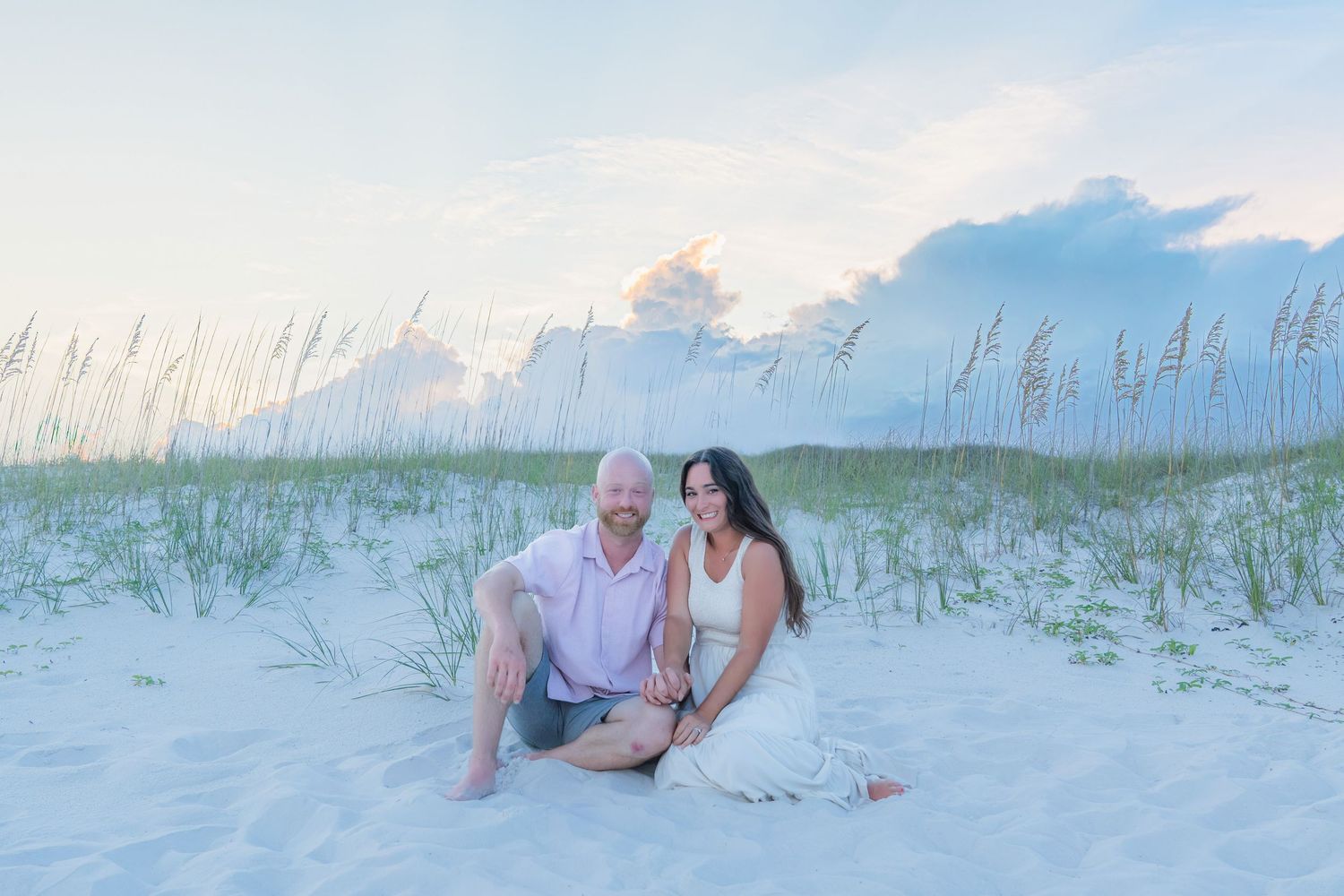 Couple sitting together on white sand beach dunes with sea oats and pastel cloudy sky at sunset.